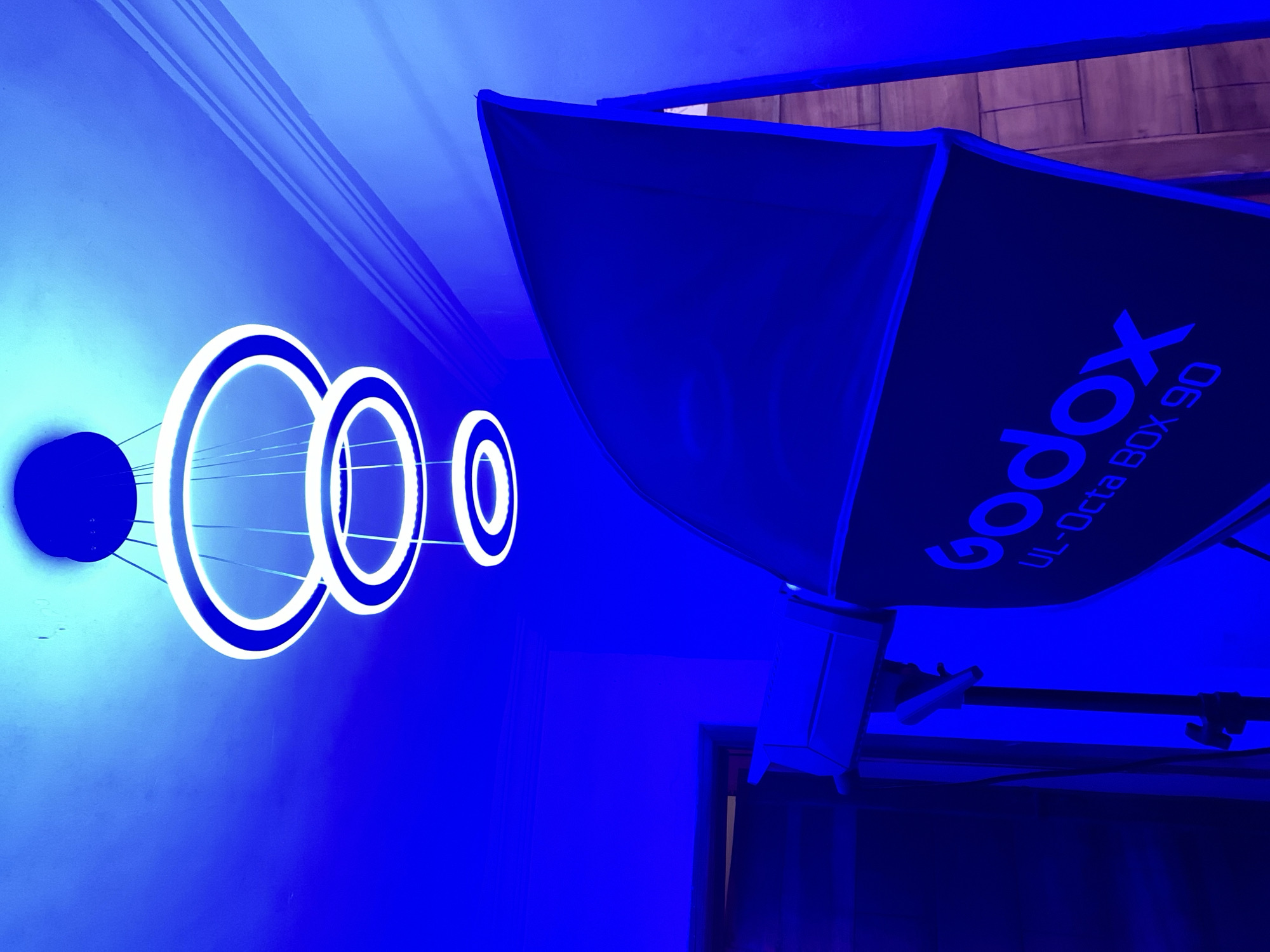 A blue-lit interior scene with a circular wall decoration featuring four connected concentric rings and a black disk. The rings are lit with a white light. To the right, there is a Godox UL-Octa BOX 90 softbox on a stand. The location is presumed to be Ikorodu, Nigeria.