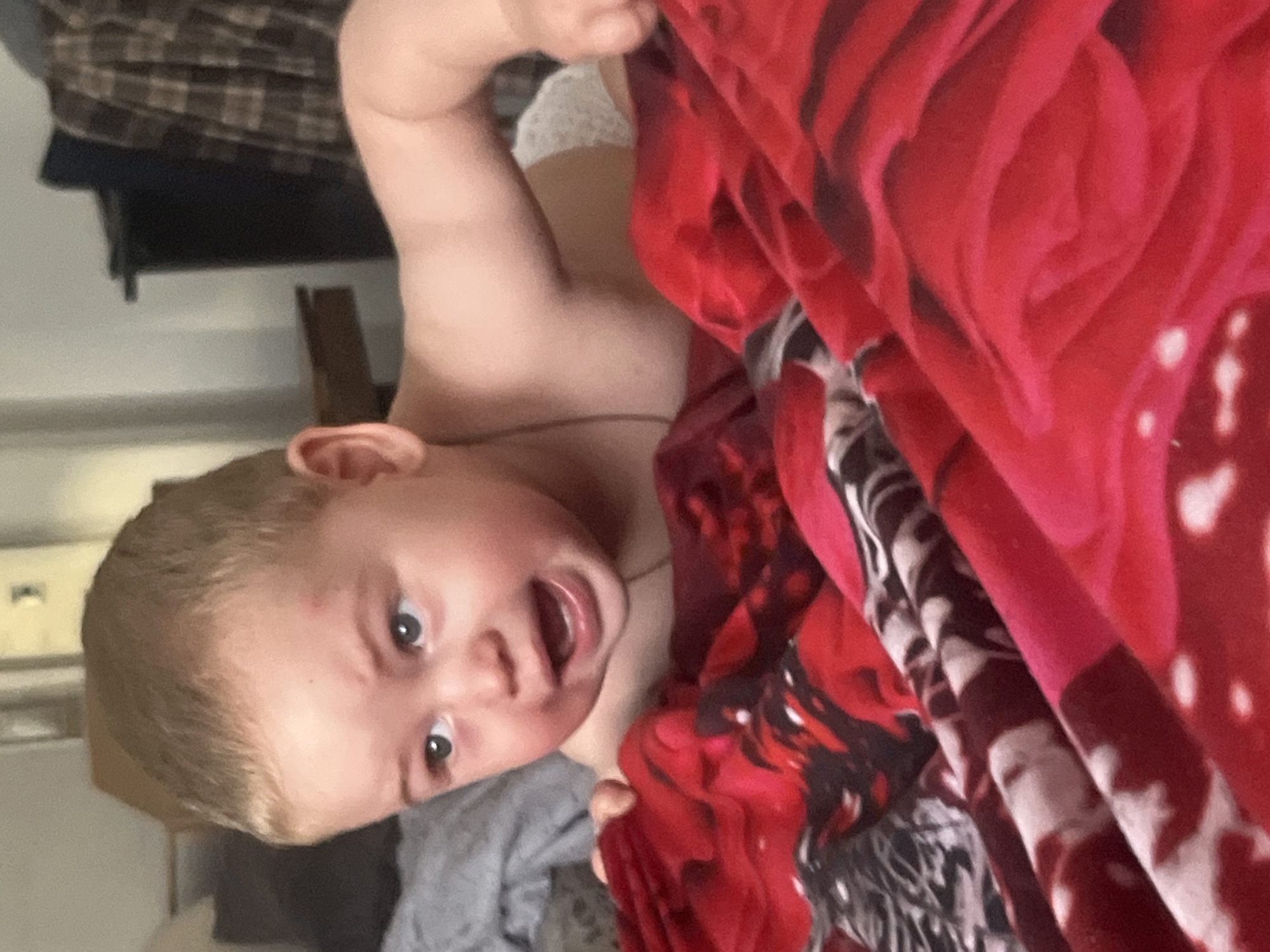 The image shows a close-up of a baby lying on a bed with a red blanket. The baby has blond hair and blue eyes, and is looking to the left with its mouth open. The blanket has a floral pattern with red and black colors. The background is blurred and appears to be a bedroom with furniture and a plaid curtain. There is no indication of the city of Chelyabinsk, Russia.