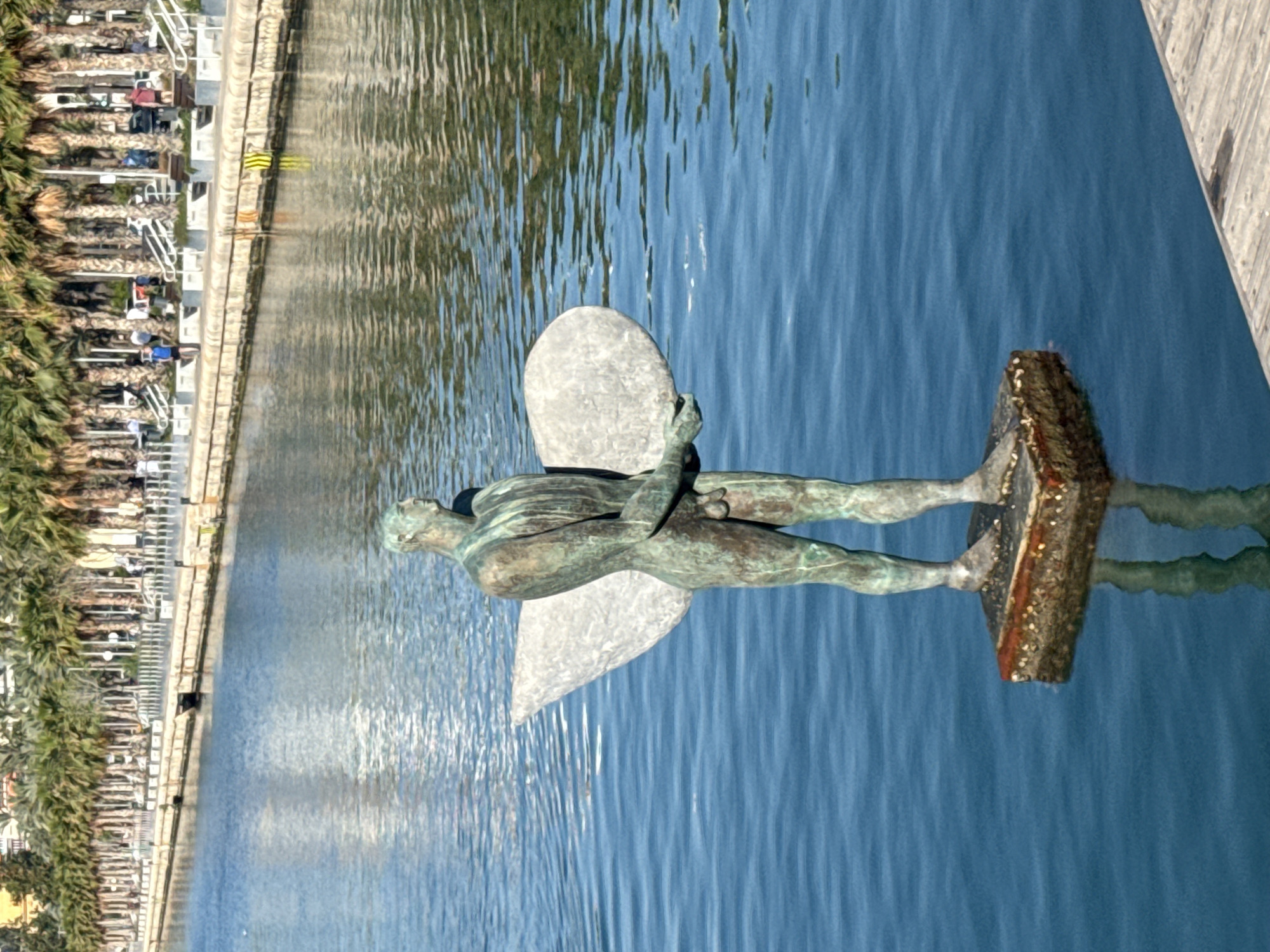 The image shows a statue of a winged figure lying horizontally on a base in the water. The statue is made of metal, possibly bronze. The background consists of a body of water, potentially a harbor or canal, with a waterfront promenade on the left side, featuring palm trees and white architectural structures. The scene appears to be located in a coastal urban environment.