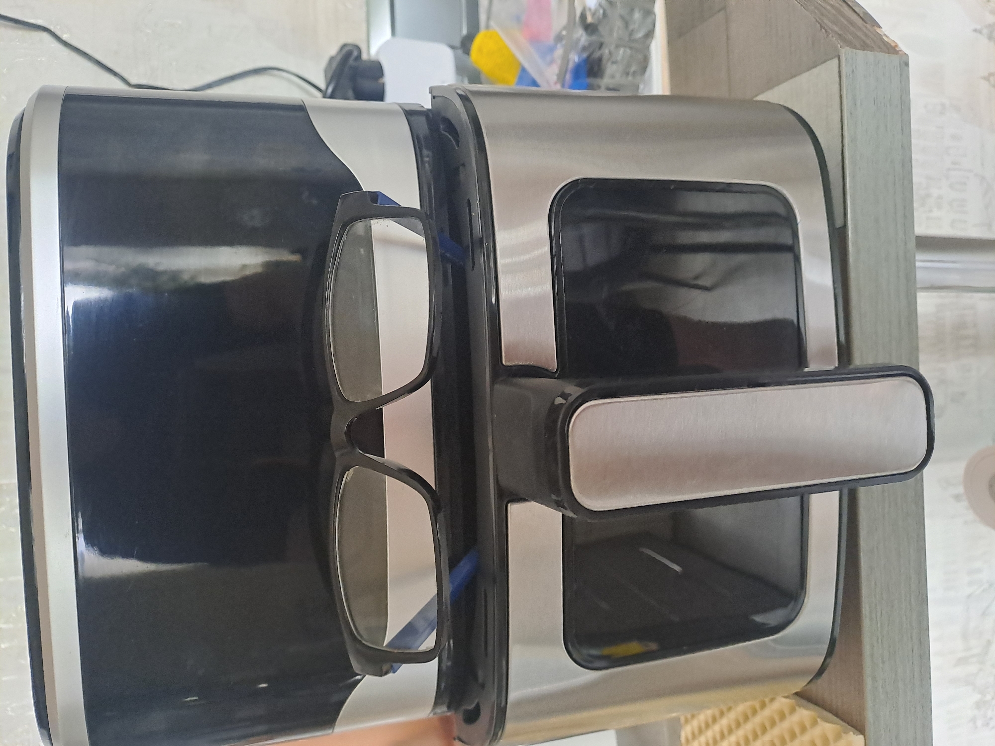 This image, originating from Elektrostal, Russia, presents an indoor scene focused on a kitchen appliance. A two-part appliance, identified as a cooking device, likely an air fryer, occupies the central frame. Its lower section is constructed from brushed stainless steel with a prominent black plastic handle and a dark, transparent panel; the upper section is composed of glossy black plastic. A pair of black-framed eyeglasses with clear lenses rests horizontally on the top edge of the lower section, leaning against the front of the upper section. The appliance is positioned on a light grey-toned horizontal surface, appearing to be a countertop or shelf. In the out-of-focus background to the right, a yellow object, various clear plastic items, and a black electrical cord are discernible. The wall behind features a light-toned, subtly patterned surface. No individuals or active interactions are visible within the frame.