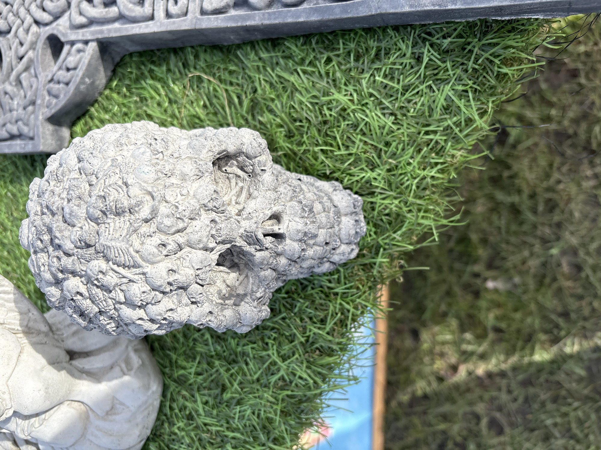 A high-angle shot shows an ornamental skull, sculpted from a gray material, seemingly stone, placed on artificial green grass. The skull's surface is textured with small skull-like shapes, indicating a possible theme of mortality. Adjacent to the skull is a dark gray decorative architectural element, possibly part of a larger structure. Beneath the artificial grass, a patch of blue and brown is visible.
Without additional context, it's impossible to confirm the location specifically in Chirk, United Kingdom. However, the image seems to depict a garden or decorative setting.
