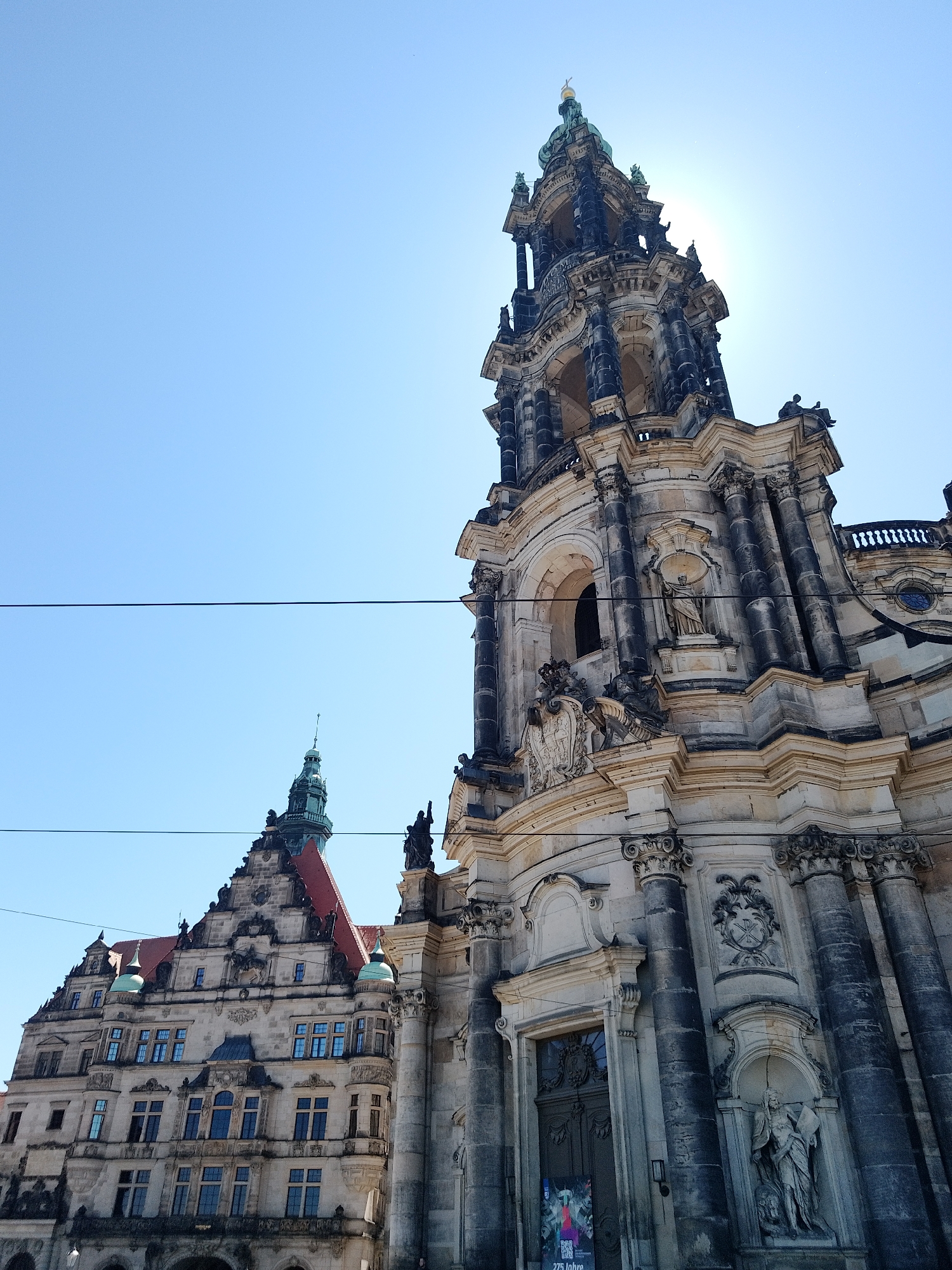 The image shows two buildings in Dresden, Germany, under a clear blue sky. On the right is the Hofkirche (Dresden Cathedral), a baroque-style Catholic church with an ornate tower and exterior sculptures. To the left is another building with a red-tiled roof and a smaller, pointed tower, also showing baroque architectural details. The perspective is from below, looking upwards, emphasizing the height of the structures. A set of two electrical wires bisect the sky.
