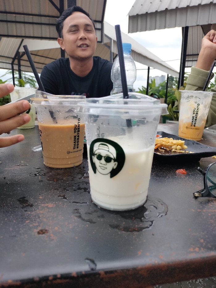 The image depicts a casual outdoor or semi-outdoor gathering, likely at a cafe or food stall in Dramaga, Indonesia, under an overcast daytime sky. In the background, a man with dark hair, wearing a black t-shirt, is seated and looking forward with his eyes closed or squinting, appearing relaxed. Fragments of other people are visible: a hand on the left near a faint green drink cup, and an arm on the right holding a yellowish-orange drink.
The foreground is dominated by a dark, possibly metal or distressed wooden table, visibly wet with condensation rings and spills from drinks. Multiple clear plastic cups with black straws are arranged on the table. A prominent cup in the center holds a white, milky beverage and features a circular green logo depicting a cartoon face wearing a baseball cap and sunglasses. To its left, another cup contains a light brown, coffee-like drink. Several cups, including the white and coffee-colored ones, display the vertical text "THANK YOU #SOBATBENTO".
Other objects on the table include a clear plastic water bottle with a blue cap, a black plate containing fried food (resembling french fries) and a dollop of red sauce, and dark-rimmed glasses resting on the far right. The setting includes an open-sided structure with a white corrugated roof and visible greenery in the distant background, suggesting a laid-back, social environment.