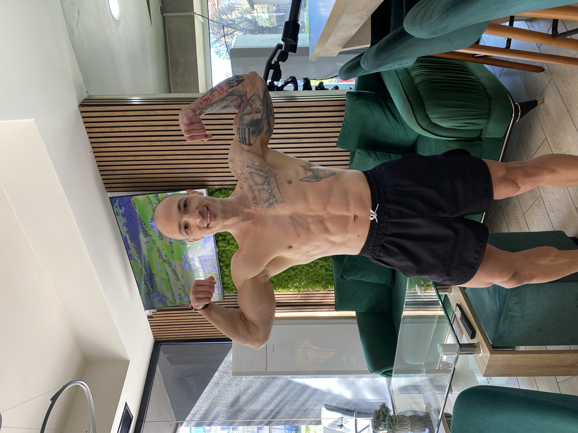 A shirtless man with a shaved head and tattoos is posing and flexing his biceps inside a well-lit building. He is wearing black shorts with a white drawstring. The interior includes green velvet chairs, a glass-topped coffee table, a wall with horizontal wooden slats, and a large screen displaying an outdoor scene. The man is in Pleven, Bulgaria.