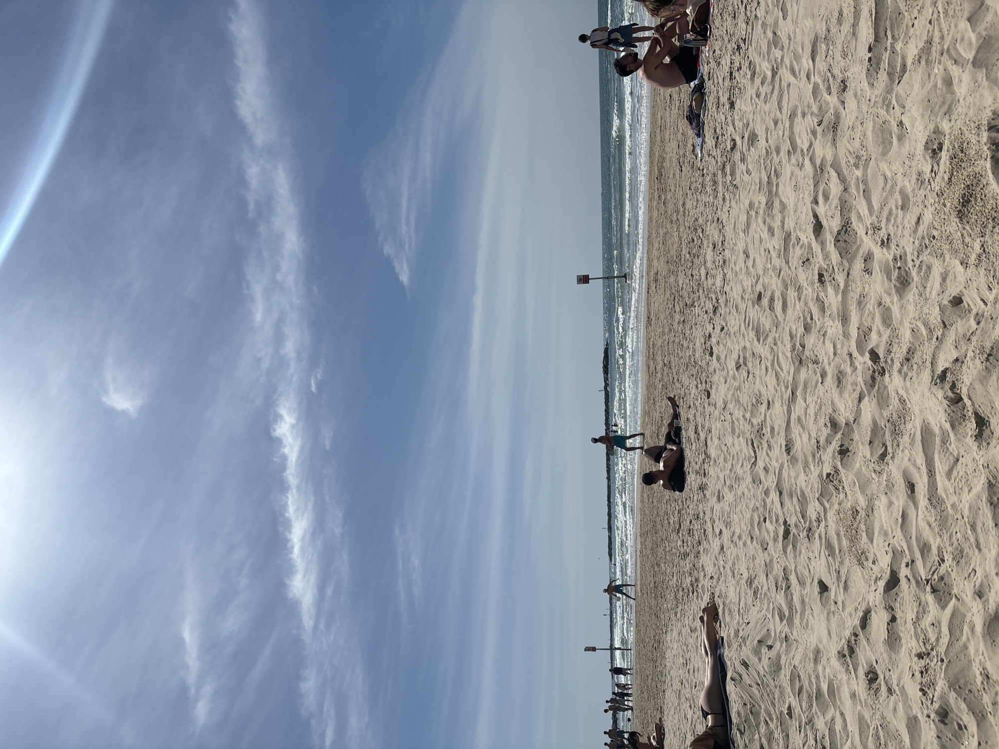 This image depicts a sunny beach scene in Tel-Aviv, Israel, under a bright blue sky with scattered cirrus clouds and a visible sun glare emanating from the upper left. The foreground consists of light-colored sand, textured with footprints and undulations.
Multiple individuals are present across the beach. In the immediate foreground left, a person in a dark swimsuit lies prone on a light-colored towel or mat. In the mid-foreground, two individuals are lying on the sand, one on their stomach, facing the water. To the right, a shirtless man with a beard sits on a colorful blanket next to another partially visible person, with various small items like bottles nearby.
Closer to the waterline, several individuals are engaged in activities. Two people are playing Matkot (beach paddleball) at the water's edge. Another person in light blue shorts stands in the shallow water, facing the beach. A person in light attire stands further right, facing the ocean. A cluster of approximately five to seven individuals is visible further down the beach to the left, near the waterline.
The Mediterranean Sea exhibits moderate waves with foamy whitecaps. Two vertical poles stand in the shallow water; one features a red rectangular sign with a white circle and a diagonal line, indicating a restriction. In the distant background, several dark breakwater structures extend into the sea. The scene portrays a lively recreational beach environment.