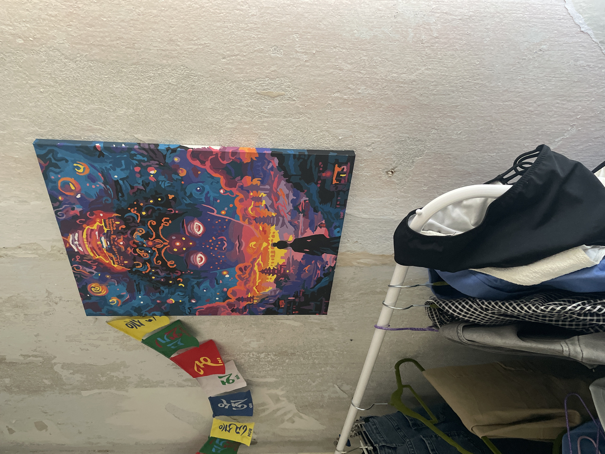 An indoor scene shows a wall with a painting and prayer flags. The painting depicts a cosmic landscape with a large, stylized face. Below the painting, a clothing rack holds various garments. To the left of the painting, a string of colorful prayer flags hangs against the textured wall. The wall appears to be made of concrete with peeling paint. The overall environment suggests a domestic setting. The location of Kopeysk, Russia is not determinable from the visual evidence.