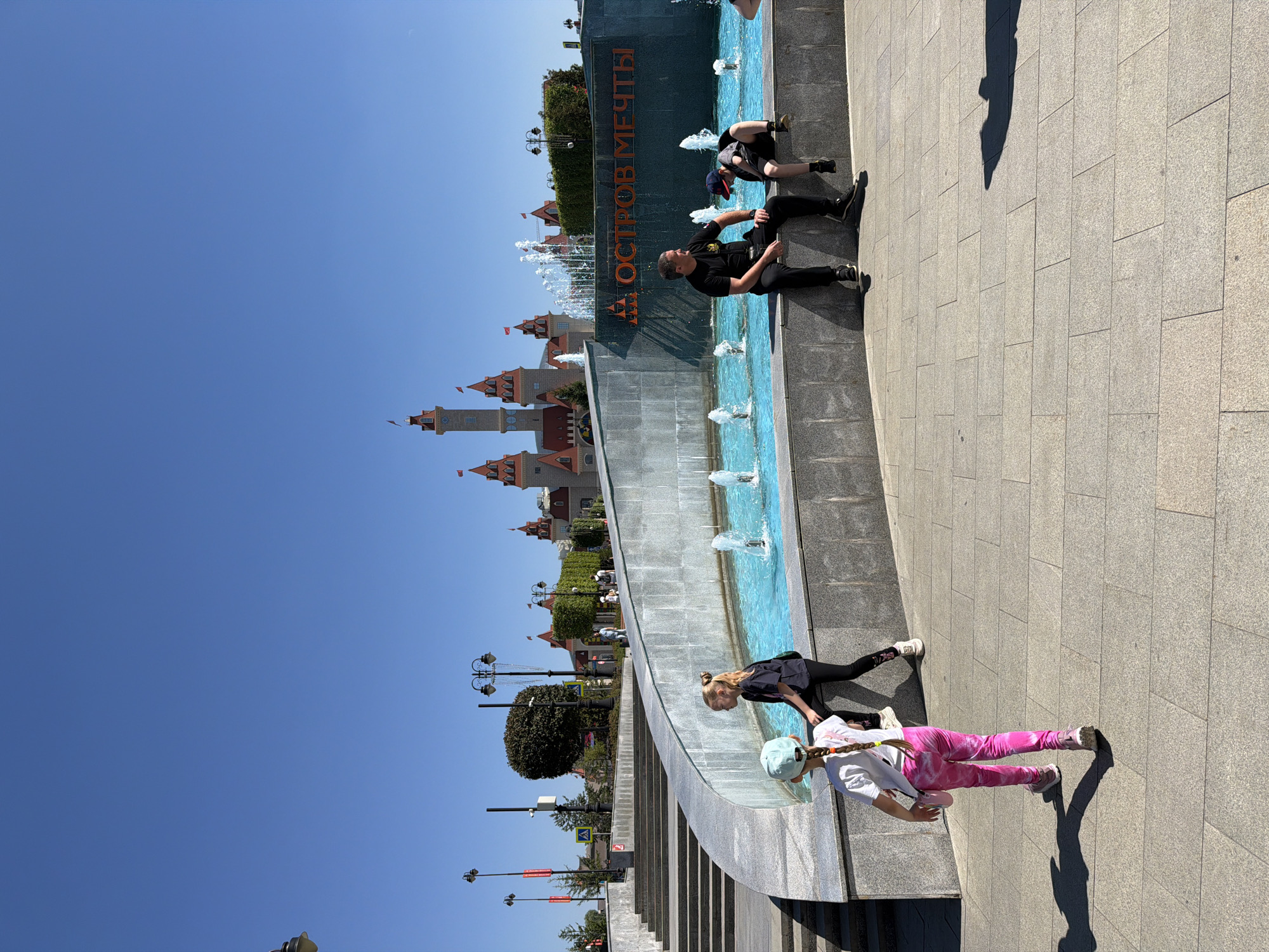 This image depicts a sunny day at the "Dream Island" amusement park in Moscow, Russia, specifically in front of a large fountain. In the foreground, two young girls are walking on a paved walkway, with the girl on the left wearing pink leggings and a light blue hat. To the right, a man in a black t-shirt and pants is sitting on the edge of the fountain, facing a young boy who is also sitting and appears to be looking down. The fountain features bright blue water and several spray jets. In the background, a large, castle-like structure with red roofs and flags is visible, and a sign with orange Cyrillic letters spelling "ОСТРОВ МЕЧТЫ" (Dream Island) is mounted on the fountain's wall. The sky is a clear, bright blue.