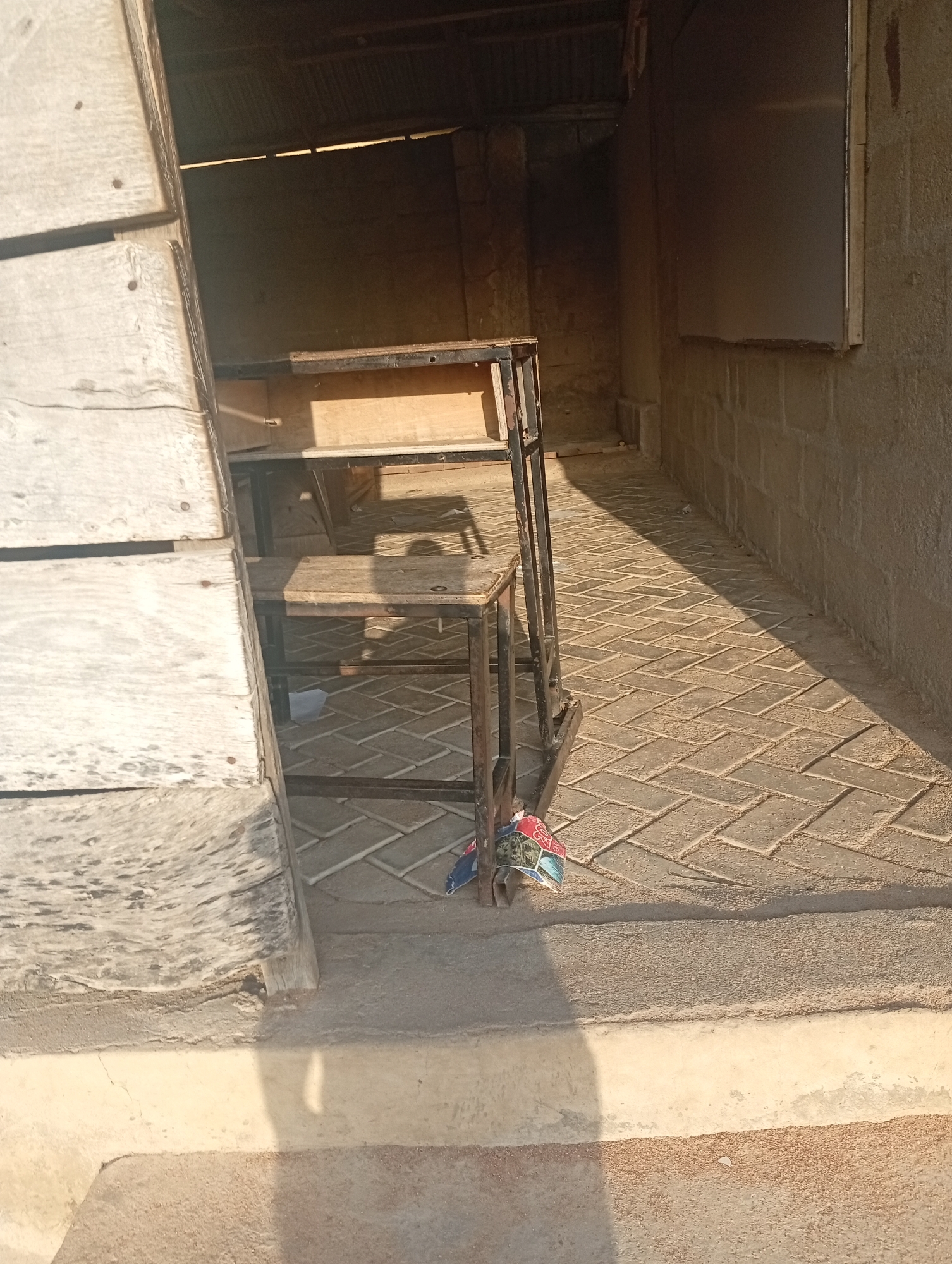 The image depicts an outdoor scene, possibly a shop or stall, in Jalingo, Nigeria. In the foreground, a portion of a wooden structure is visible on the left, with weathered planks. To the right, a simple metal and wooden shelf unit is positioned. The shelves are tiered and appear to be made of wood with a metal frame. At the base of the shelf unit, a small bundle of colorful items, possibly packaging or fabric, is attached. The ground is paved with interlocking bricks laid in a herringbone pattern. A distinct shadow stretches across the brickwork from the shelf unit. Behind the shelf unit, a wall constructed of concrete blocks is visible, and further back, a corrugated metal roof is suggested by the darkness and texture. The overall impression is one of a functional, perhaps somewhat rudimentary, commercial space in an urban environment.