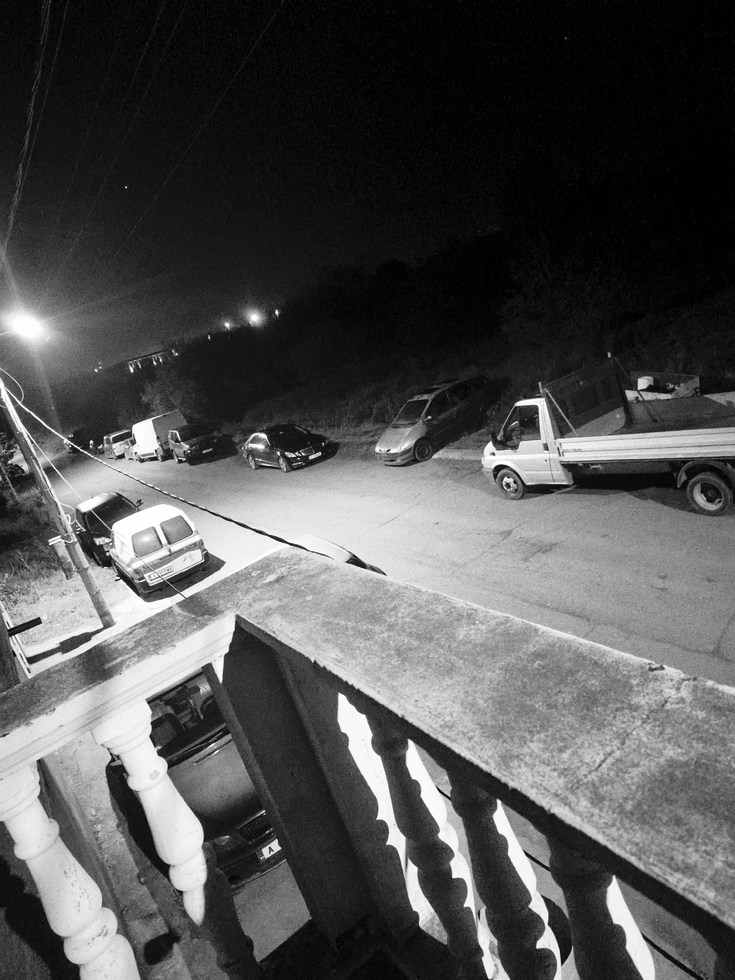 This black and white photograph captures a quiet street scene at night in Burgas, Bulgaria, viewed from a slightly elevated position, possibly a balcony or terrace, indicated by the balustrade in the foreground.
The primary subjects are various vehicles parked along a two-lane asphalt road. On the right side, a white Ford Transit pickup truck with an open bed is prominently displayed, followed by a silver hatchback, and further down, a black sedan. On the left side of the street, closer to the viewer, is a white van with rear doors, and a dark-colored car partially obscured behind it. Other indistinct vehicles and what appear to be utility poles with overhead lines line the left side of the road.
There is no specific human activity visible; the scene depicts parked vehicles under the glow of streetlights. The time is clearly night, as evidenced by the dark sky, the illuminated streetlights casting long, stark shadows, and the general low-light conditions. A single bright star or planet is visible in the upper left portion of the sky. The weather appears clear.
Notable details include the coarse concrete texture of the balustrade railing in the foreground, with pronounced shadows from its balusters. Streetlights create strong highlights and deep shadows across the road and vehicles. Visible text includes a Bulgarian license plate "A 9801 MA" on the rear of the white van, and the letter "A" of another license plate on the black sedan. The word "TRANSIT" is faintly visible on the side of the white pickup truck. In the distant background, more lights suggest additional structures or a continuation of the street on higher ground, with hills or dense foliage outlining the horizon.