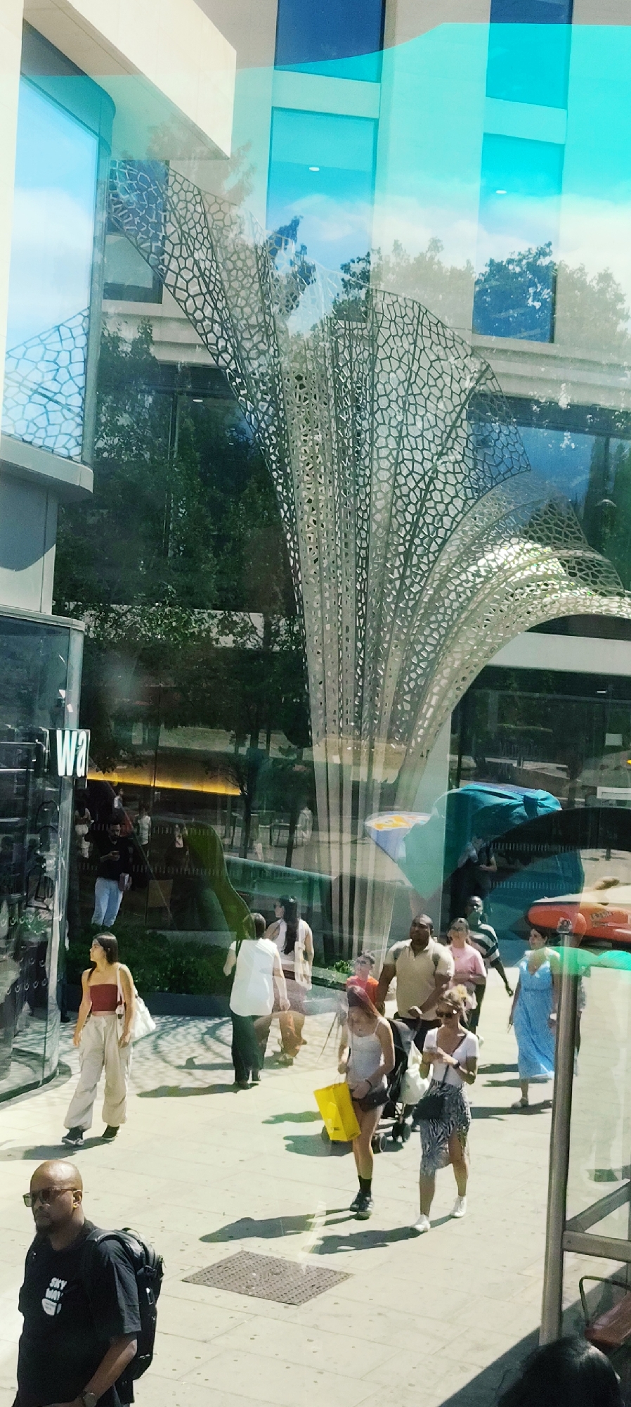 This is a street-level, eye-level shot of a busy pedestrian area, likely in a modern urban environment, possibly in London, United Kingdom, given the architectural style and blue glass elements common in some city buildings. The scene is dominated by reflections on a large glass facade, which overlays the actual street activity.
In the foreground, a bald Black man with sunglasses and a black t-shirt with white lettering, carrying a black backpack, is looking to his left. To his right, a group of pedestrians walks away from the viewer. Among them is a woman in a white top and a patterned skirt, holding a phone, and a younger woman in a white tank top and dark shorts, carrying a bright yellow shopping bag and wearing black socks and shoes. Behind them, a man in a light brown t-shirt pushes a stroller. Further back, a woman in a blue dress walks towards the right of the frame. Other individuals are visible in various states of motion, including a person in a white t-shirt and dark pants, and a woman in a red tube top and light-colored pants with a white bag.
The background is heavily influenced by reflections. The glass facade shows the sky, trees, and parts of buildings. A distinctive, large, metallic sculpture with an intricate, web-like pattern is prominently featured, appearing as if it's part of the environment or a reflection. The overall impression is one of a vibrant public space with a mix of people and modern architectural elements, partially obscured by the reflective nature of the glass.