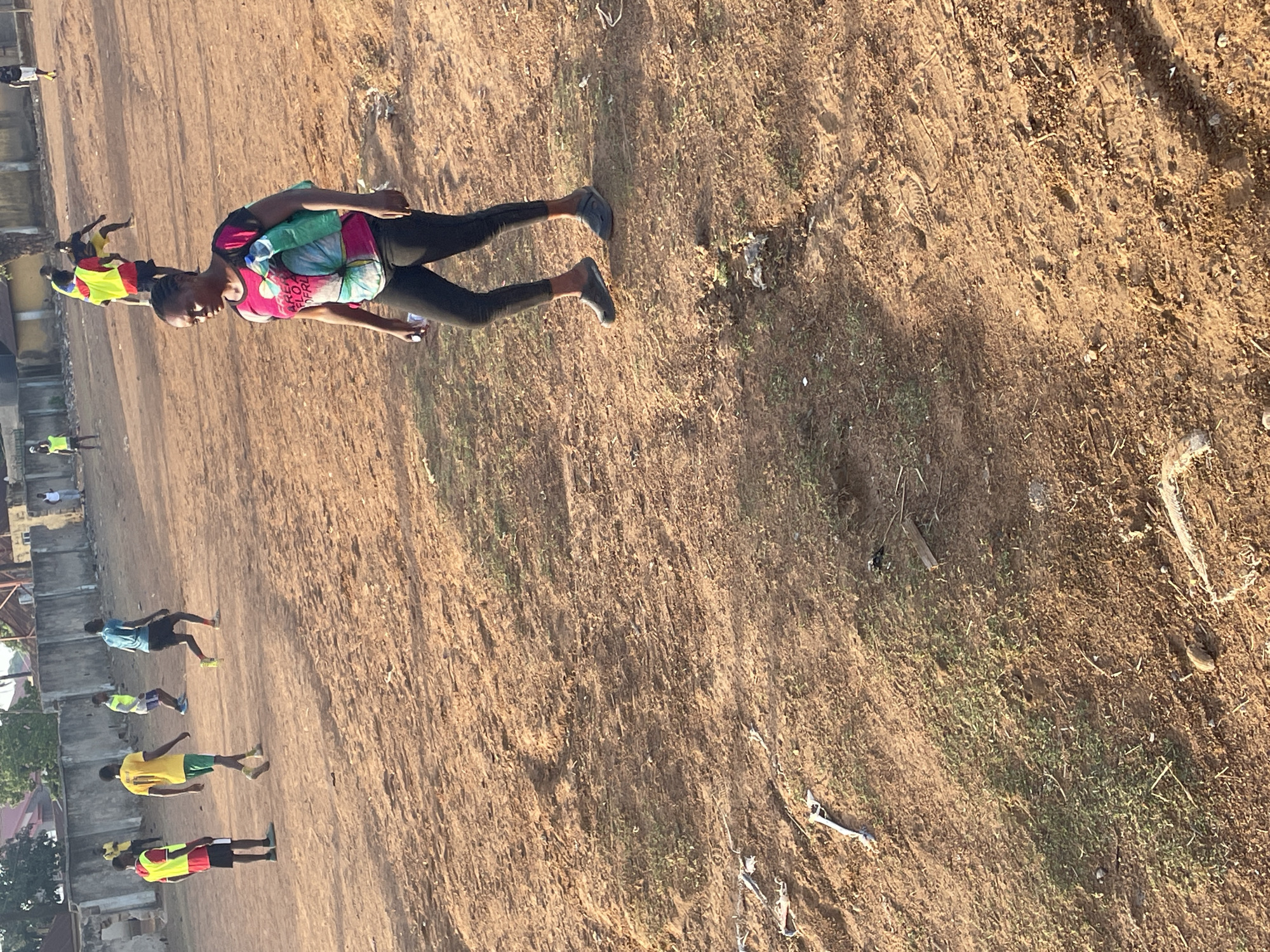 The image shows a group of people participating in an activity, likely a race or training exercise, on a dirt field in Akwanga, Nigeria. The field appears to be located near a residential area, with buildings visible in the background. Several individuals are running, wearing athletic clothing and some with brightly colored vests. One woman in the foreground is wearing a pink shirt, black leggings, and has a backpack. The ground is dry and dusty with patches of grass and scattered debris.