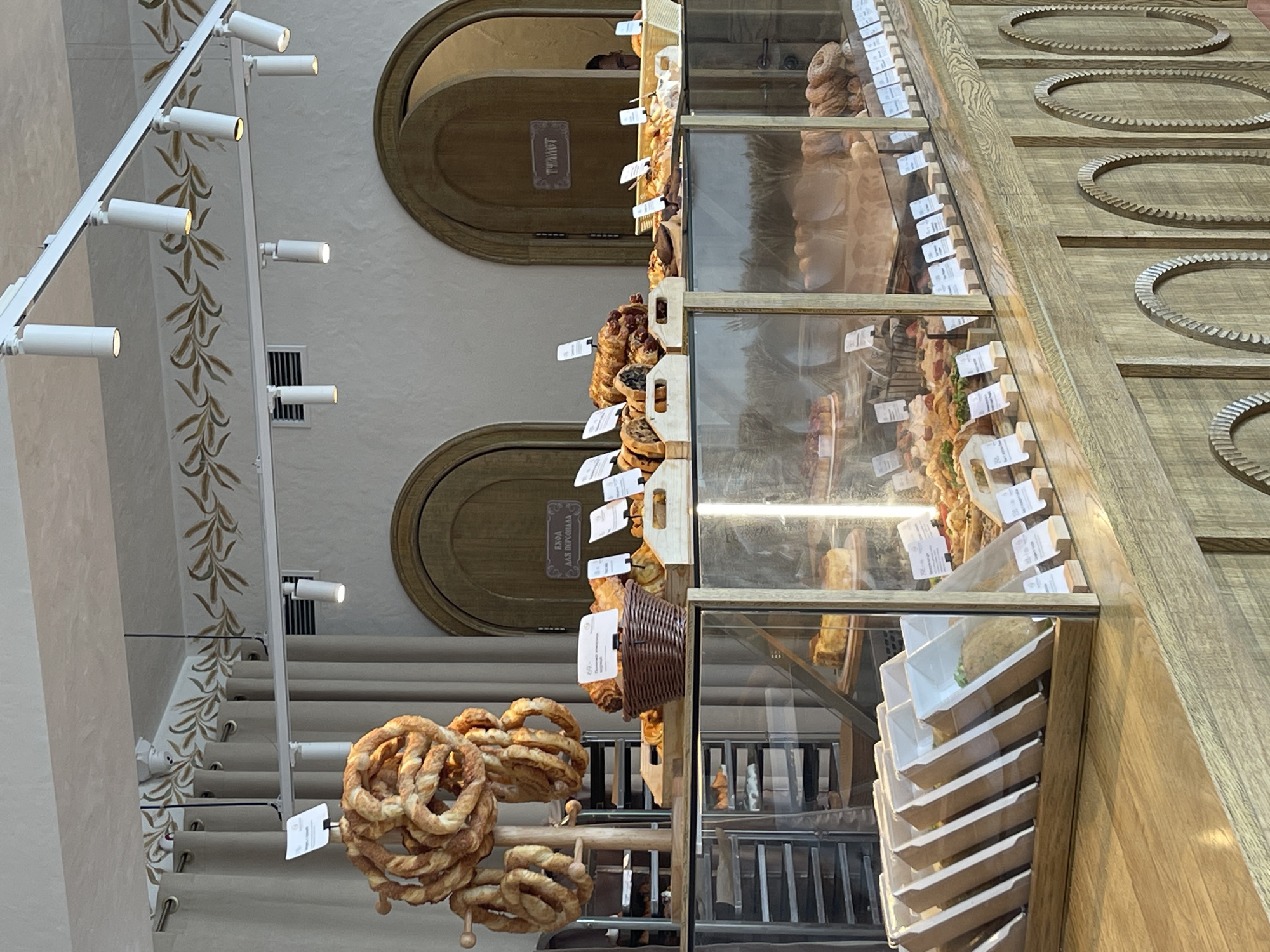 The image shows the interior of a bakery. The scene is dominated by a display counter filled with various baked goods, including pretzels, croissants, bread, and pastries. The display is made of glass and wood. Behind the counter, there are arched wooden doors. The walls are decorated with a leaf motif. There are also several light fixtures visible. The overall atmosphere appears to be bright and inviting. The location is not specified beyond "Unknown, Unknown".