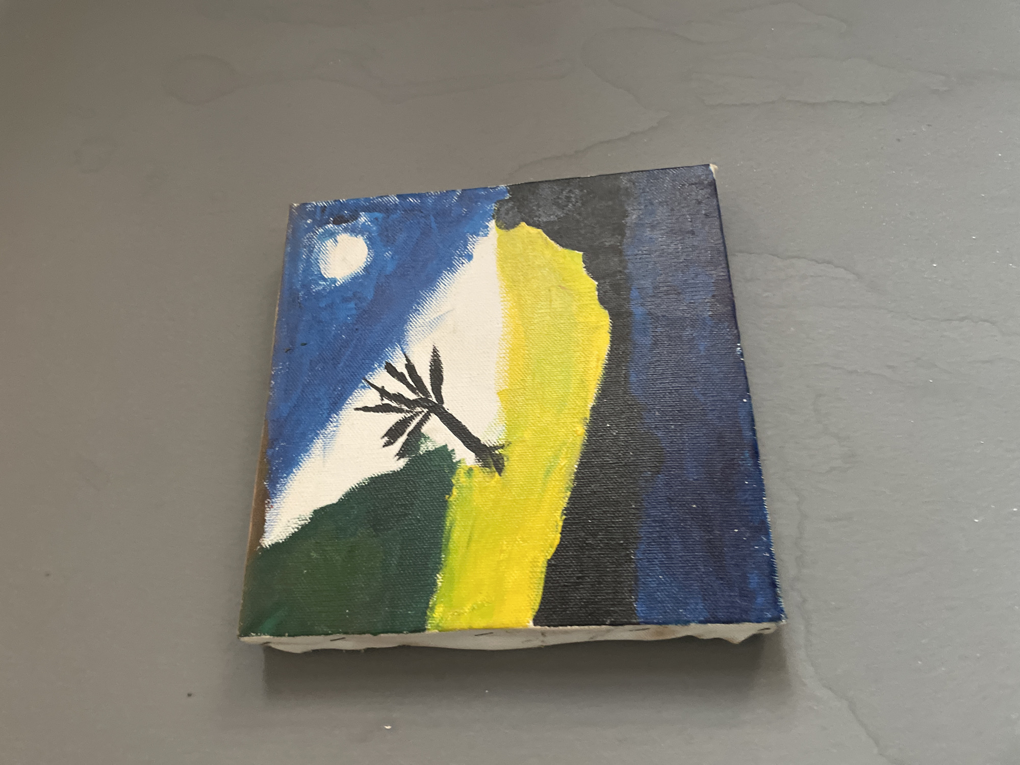 The image shows a small, amateurish oil painting on canvas. The painting depicts a landscape scene with a stylized tree in the foreground. The sky is blue with a white moon. The ground is green on one side, separated by a bright yellow area from a dark blue section. The painting's edges are rough, indicating it may be a study or practice piece. The painting is placed on a gray surface, likely a tabletop. No information about Kurunduma, Nigeria can be ascertained from this image.
