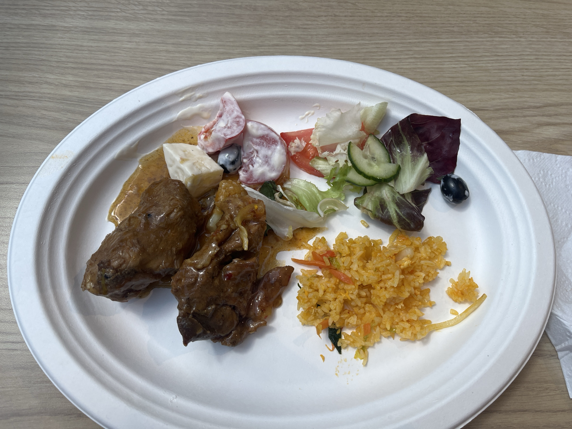 The image displays a white, round plate with a textured rim, filled with a meal. The plate is positioned on a light brown, wood-grained surface, suggesting a table. The food on the plate consists of several components. On the left side, there are two pieces of dark brown meat covered in a glistening sauce. Adjacent to the meat, towards the center-top of the plate, is a portion of cubed white cheese, with small pieces of tomato and a single black olive scattered around it. To the right of the meat and cheese, there is a small salad with lettuce, cucumber slices, and pieces of tomato. Further to the right, a pile of orange-colored fried rice is visible, with flecks of orange carrot and some green vegetables mixed in. A single strand of something, possibly fried potato, extends from the rice. The lighting appears to be from above, casting a slight shadow. The overall impression is a casual meal served on disposable tableware. There is no visible text in the image.