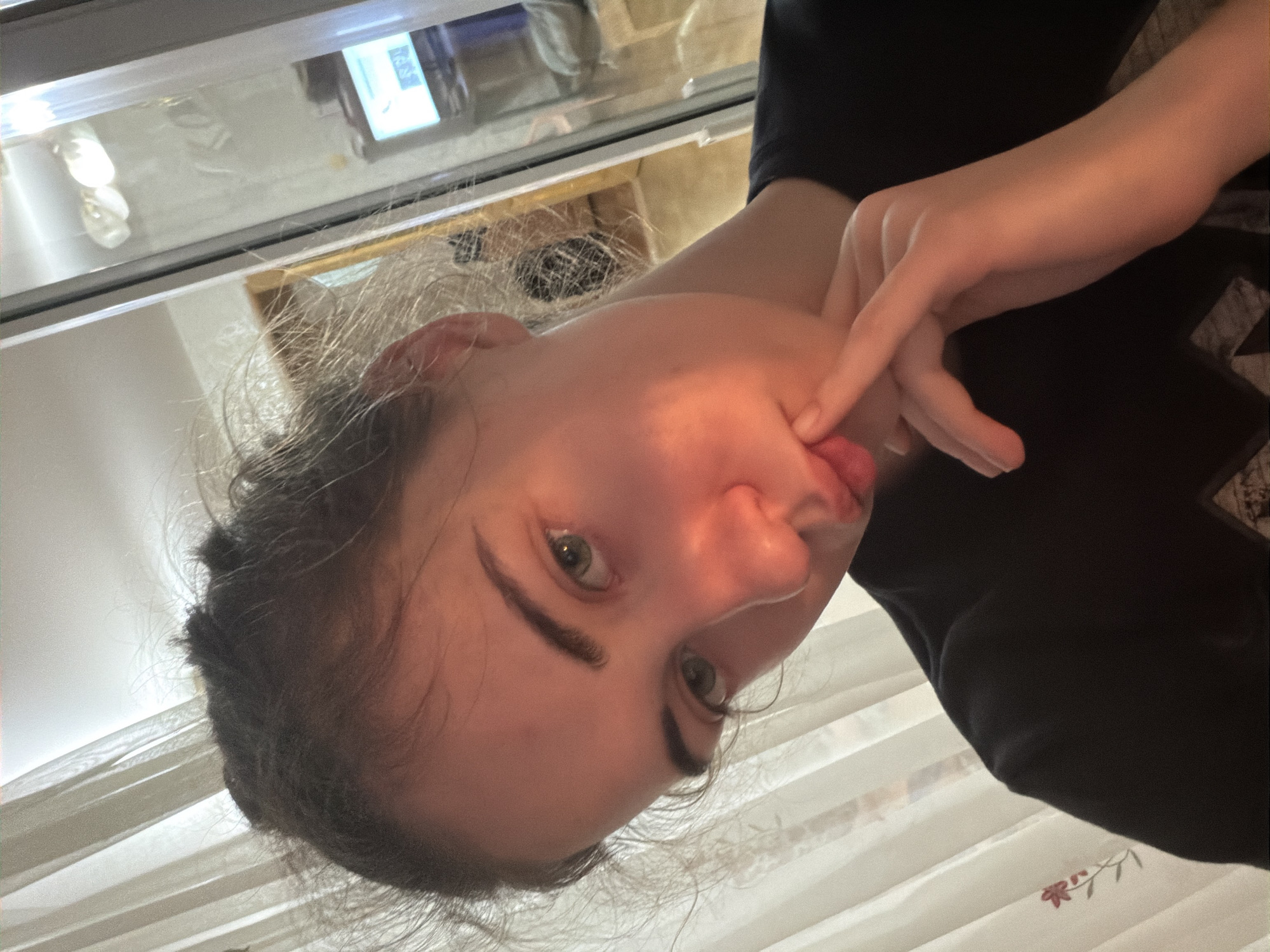A young woman with light skin and medium brown hair pulled back in a messy bun is looking into the camera. She has light green eyes and is wearing a black t-shirt. Her right hand is raised to her face, with her index finger touching her lips. Sunlight is casting a warm glow on her face, highlighting her features and creating shadows. She is indoors, with sheer curtains in the background to her left and a glass door or window to her right, through which a dimly lit room with a television and some objects can be seen. The scene appears to be in a residential setting, possibly a home in Zavodoukovsk, Russia, given the context.
