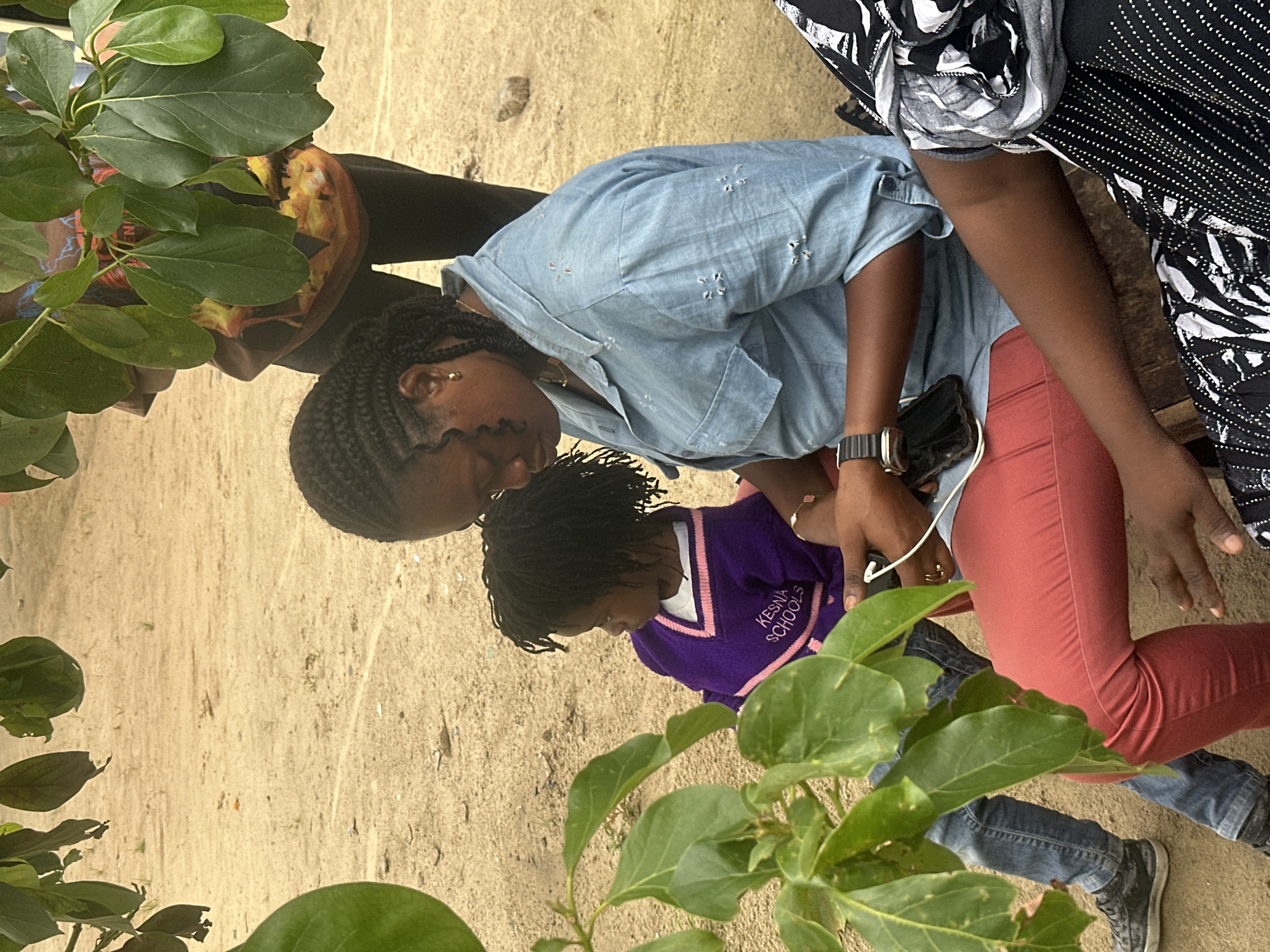 A medium shot captures three individuals seated on a wooden bench in Ganyingbo, Nigeria. In the foreground, a young child wearing a purple sweater with pink trim that reads "KESNA SCHOOLS" sits to the left, looking to the right. To the child's right, an adult woman in a light blue denim shirt and red pants is looking down at a smartphone in her hands. To the far right, another person is partially visible, dressed in a black and white patterned outfit. The ground is sandy with scattered small stones and sparse vegetation. Overhead, large green leaves from a tree obscure parts of the background. The scene appears to be outdoors, possibly in a schoolyard or a casual gathering area.