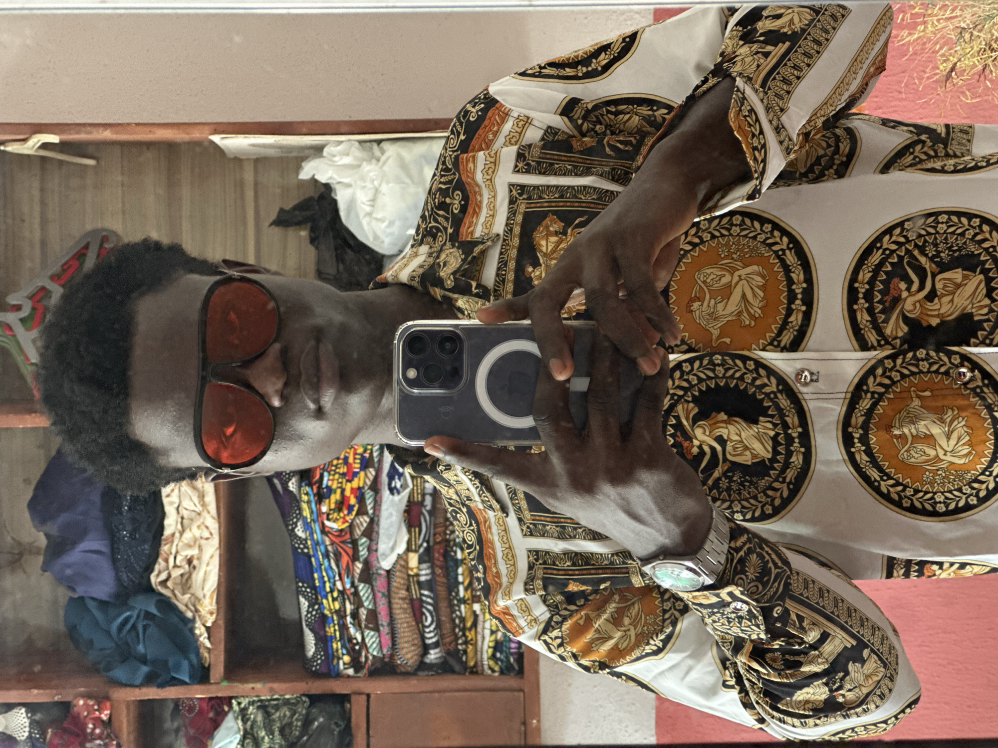 A person is seen in a selfie taken in front of a mirror inside a structure, possibly a dwelling, in Badagry, Nigeria. The person wears red-tinted glasses and a patterned shirt with medallions and ornamental designs in gold, black, and white. They are holding a smartphone, an iPhone, which obscures part of their face. A silver wristwatch is visible on their left wrist. The background contains shelves filled with folded fabric in various colors and patterns, with some white and black material on a shelf near the top. The room has a pink or peach-colored wall on one side of the mirror and a wooden frame around the shelves.