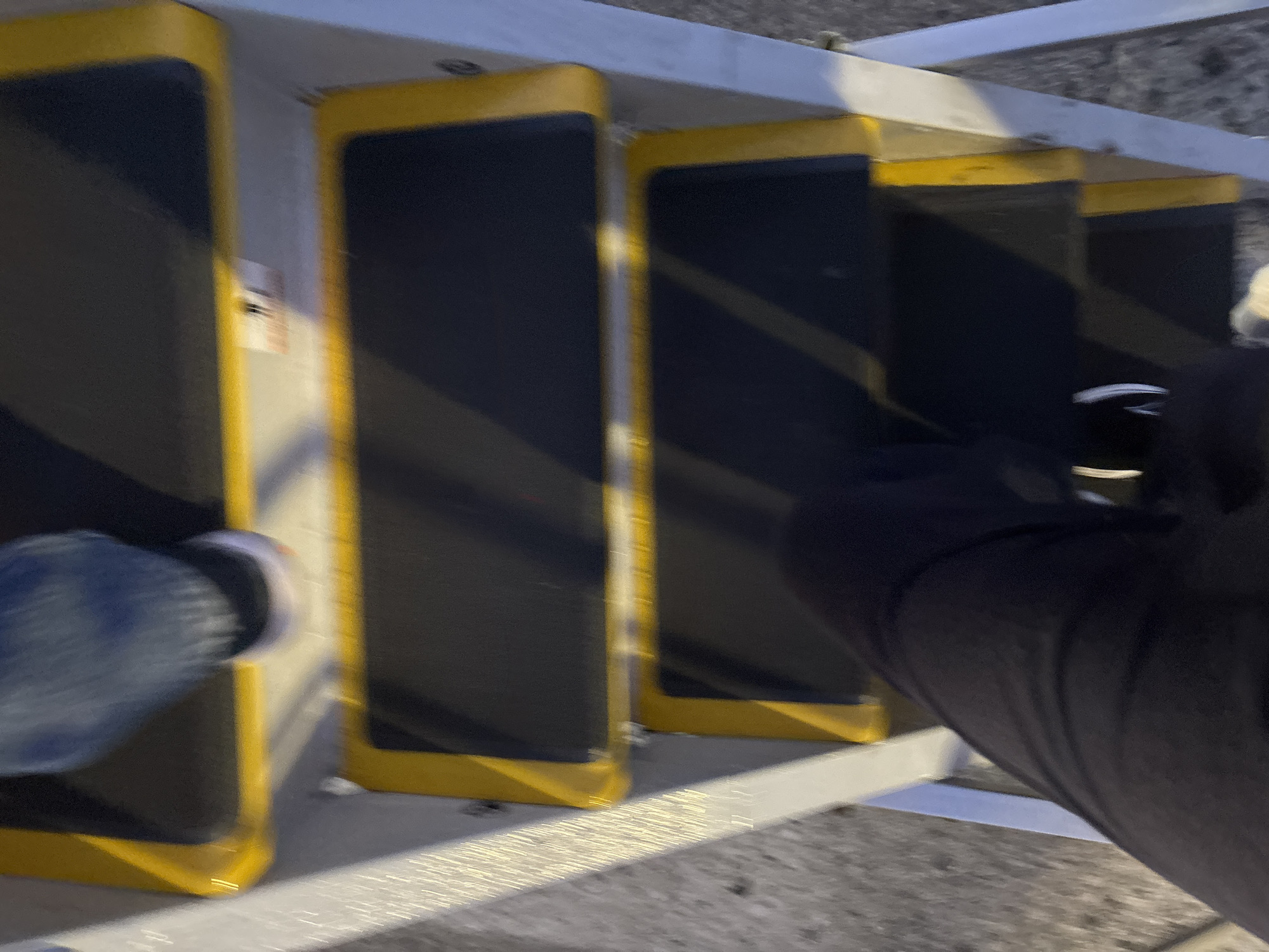 The image shows a close-up view of a person's legs and feet as they ascend a set of outdoor stairs, likely at the Orio al Serio International Airport (BGY) in Orio al Serio, Italy. The stairs have dark treads with bright yellow safety edges. The person is wearing dark pants and light-colored shoes. The background is blurred, suggesting movement and a focus on the act of climbing the stairs. The overall impression is one of transit and travel.