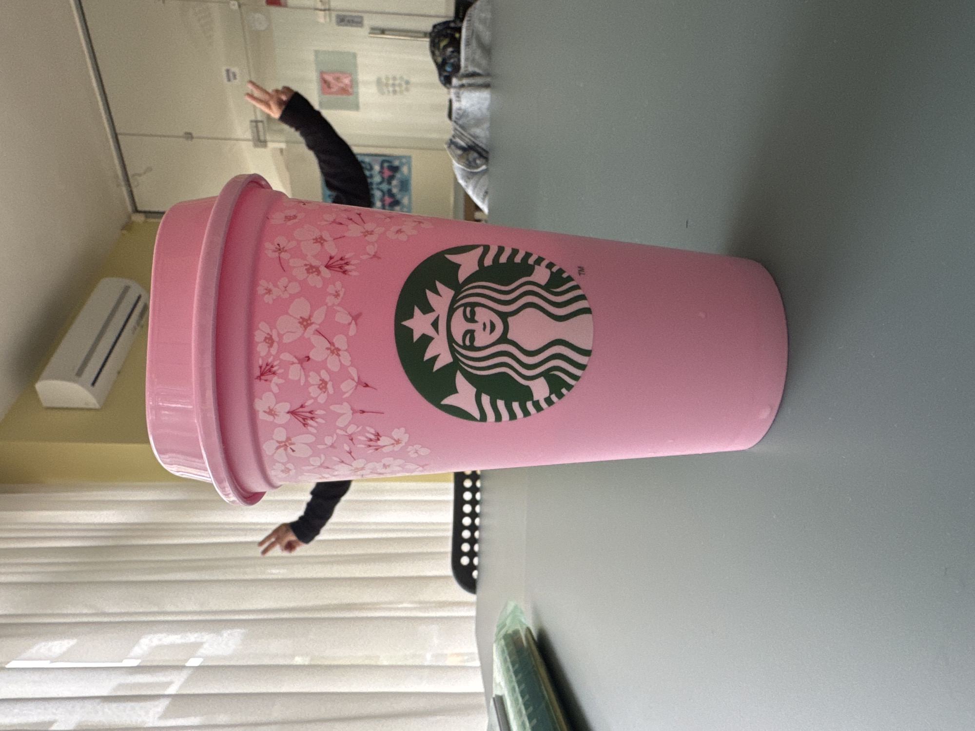 A horizontally oriented photograph captures a vibrant pink Starbucks reusable cold cup, complete with a matching lid, resting on a flat, light grey table surface. The cup is decorated with delicate white cherry blossom designs and prominently displays the iconic green and white Starbucks siren logo, accompanied by a small "TM" trademark symbol.
In the background, partially obscured by the cup, a person's arm wearing a dark long-sleeved top is visible, with their hand making a "peace" sign (V-sign). The indoor setting features white horizontal window blinds or curtains, through which natural light appears to enter. An air conditioning unit is mounted on a light-colored wall to the left of the frame. Reflections in what seems to be a mirror or glass partition reveal glimpses of a room behind, including what might be another person in light-colored attire and some wall decorations or posters. A black object with circular perforations is also partially visible. The scene suggests a casual indoor environment, possibly a common room or study area in Sofia, Bulgaria, with the peace sign conveying a lighthearted mood.
