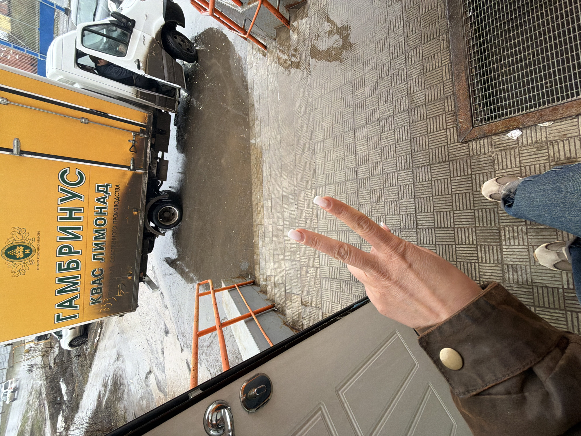 The image shows a street scene in Sarapul, Russia. A commercial delivery truck with "ГАМБРИНУС" (GAMBRINUS) and "КВАС ЛИМОНАД" (KVASS LEMONADE) written on the side is partially visible on the left. The truck is parked on a wet street next to an orange metal safety barrier. The ground is paved with gray patterned tiles and has a wet spot, possibly from recent rain. A drain grate is visible in the upper right corner. In the lower portion of the image, a hand is making a peace sign, and part of a brown jacket sleeve with a brass button is visible. The person wearing the jacket is also wearing blue jeans and gray tennis shoes. A partially-open light gray door is also visible. A man is seen inside the driver's cabin of the truck.