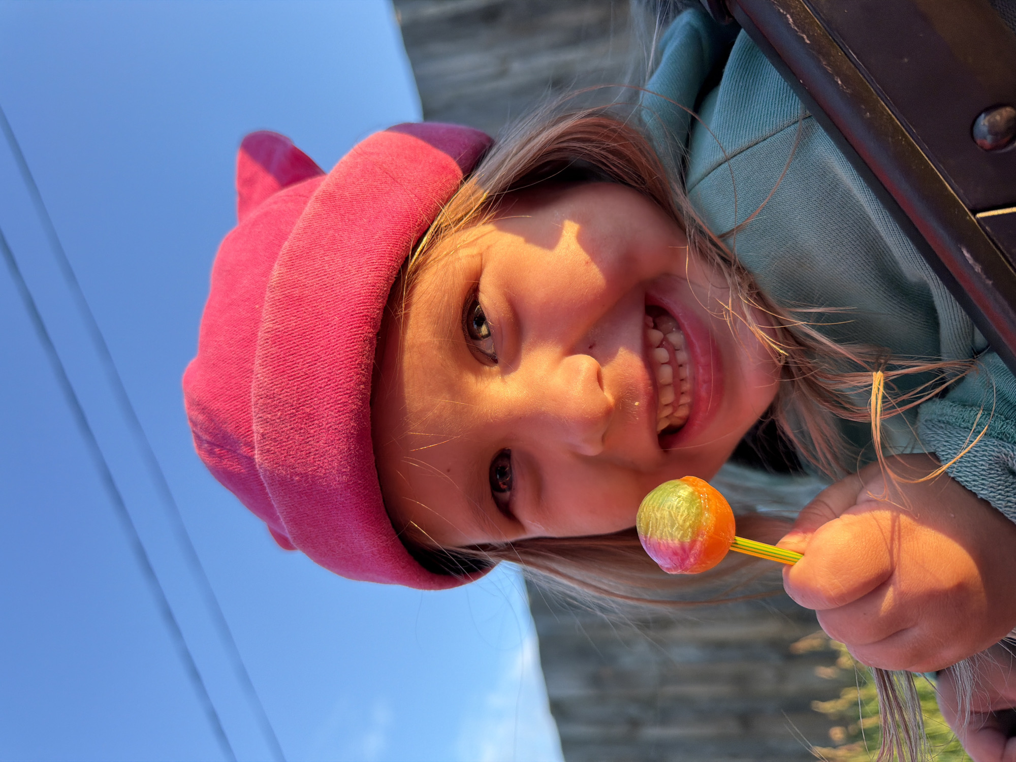 A close-up, portrait-style photograph captures a young child's happy face, rotated horizontally as if taken from a low angle. The child, with fair skin and light blonde hair, is wearing a vibrant pink beanie or hood and a teal/mint green top. She is smiling broadly, revealing her teeth, and her eyes are looking towards the viewer. Her face is warmly illuminated by golden sunlight, suggesting either early morning or late afternoon, with shadows highlighting her facial features.
In her right hand, she holds a colorful lollipop with hues of pink, yellow, green, and orange, attached to a striped yellow and green stick. The lollipop is also bathed in sunlight, giving it a soft glow. Part of a dark, possibly metallic or plastic, structure, resembling a car door frame or seat, is visible on the right side of the image, suggesting the child might be in or leaning out of a vehicle.
The background on the left is dominated by a clear, bright blue sky, indicating sunny weather. Two thin, dark lines, likely power lines or telephone wires, traverse the sky in the upper left portion. The background on the right, behind the child, is out of focus and appears to be a textured, greyish-brown surface. The overall impression is one of childlike joy and playfulness in an outdoor setting, potentially in Chaikovsky, Russia. There is no visible text in the image.