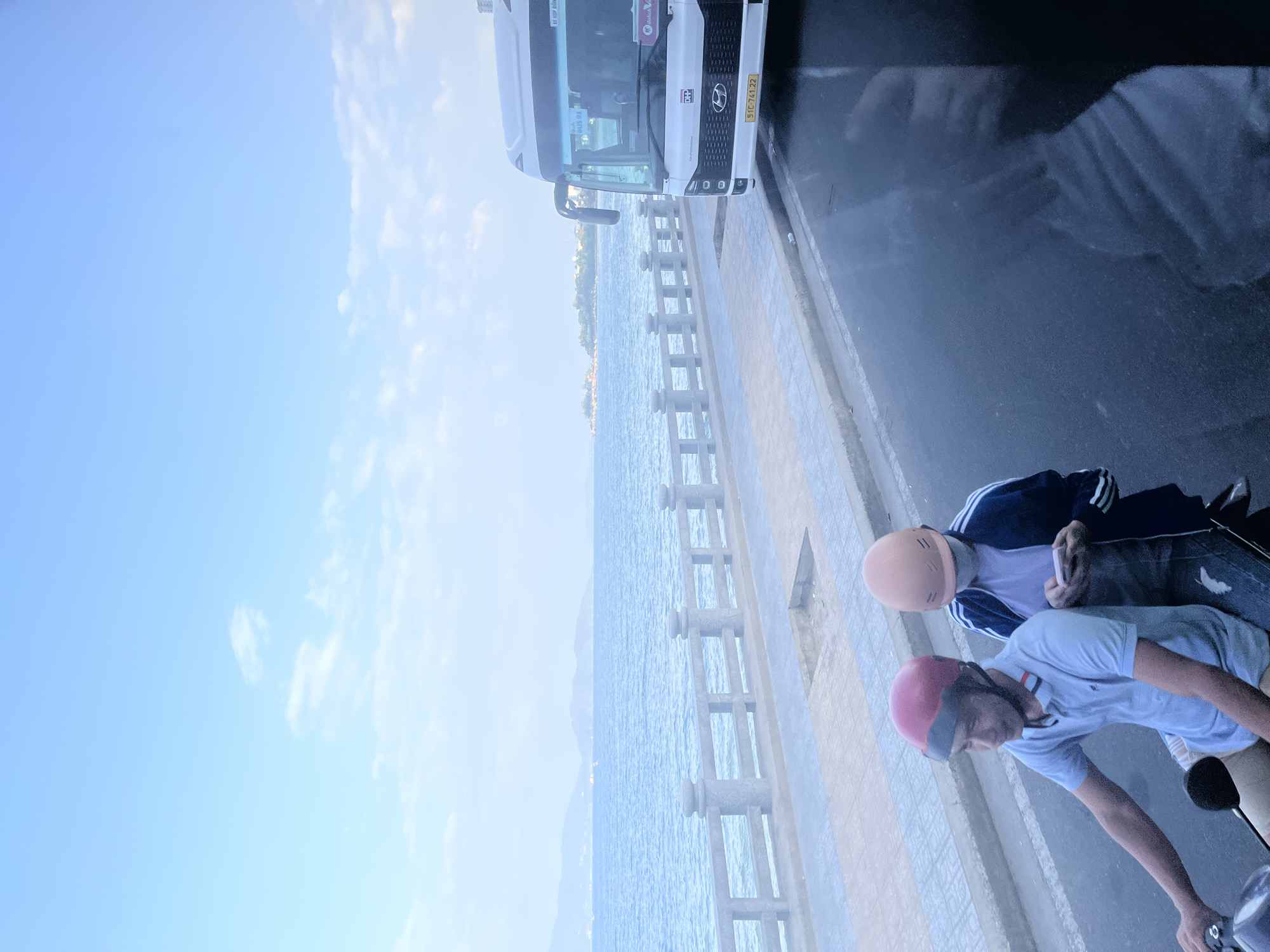 Two individuals are on a motorcycle, traveling on a wet road. The person in front, wearing a red helmet, is driving. The person behind, wearing an orange helmet and a face mask, is looking at a smartphone. To the left of the road is a sidewalk with a railing overlooking the sea. The sea is calm with a few distant islands or landmasses on the horizon. A white bus is parked on the right side of the road. The sky is clear with some light clouds. The scene appears to be in Nha Trang, Vietnam, a coastal city known for its beaches and ocean views.