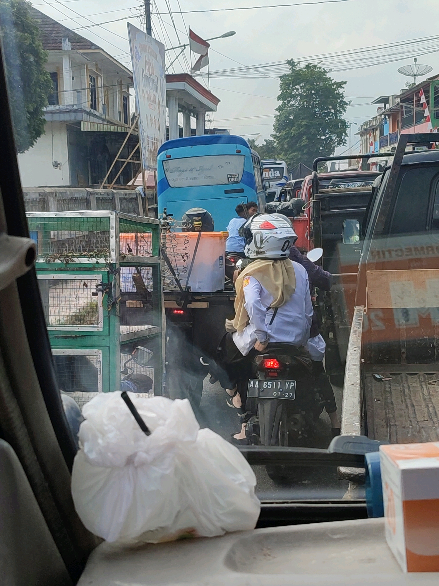 The image captures a congested street scene in Kalikajar, Indonesia, likely during daylight. The perspective is from inside a vehicle, looking forward. In the foreground, a white plastic bag with a black object poking out sits on a gray surface.
Dominating the mid-ground is a traffic jam with various vehicles. A bright blue bus is prominent, with its rear visible. To its right, a black truck with a wooden bed is stopped. In front of the truck, a motorcycle with two riders is navigating the traffic. The rider in front wears a white helmet with red and silver accents and a light brown hijab, along with a white long-sleeved shirt and dark pants. The motorcycle's license plate reads "AA 6511 YP". To the left of the bus, stacked green cages are visible, containing what appear to be animals, possibly snakes.
The background reveals a street lined with buildings, some with balconies and awnings. Red and white flags are displayed on one of the buildings. Trees with dense foliage are also present, along with overhead power lines and a satellite dish. The sky is overcast. The overall impression is one of a busy, somewhat chaotic urban environment.