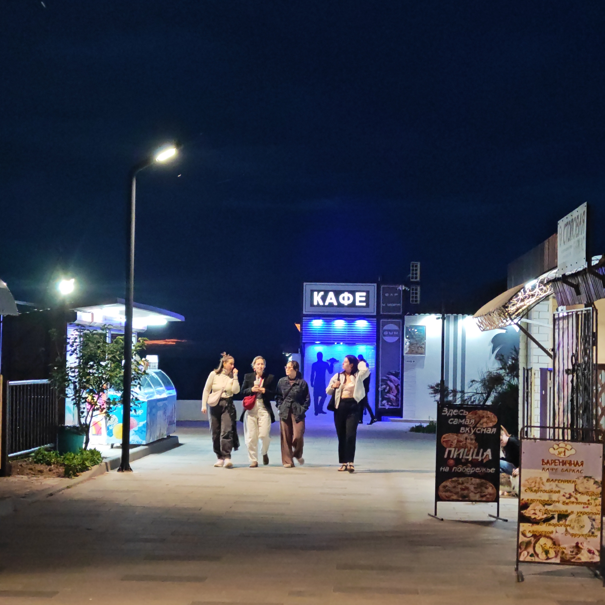 The image captures a nighttime scene on a brightly lit pedestrian promenade in городское поселение Щелкино, Ukraine. Four women are seen walking casually from left to right across the paved path. They are dressed in everyday clothing, with some carrying small bags or cups.
The area is well-lit by artificial sources, including a tall streetlamp prominently featured on the left side of the frame, and glowing signs from various establishments. The sky is dark blue, indicating the evening or night.
In the background, several kiosks and small shops are visible. A large, illuminated sign prominently displays "КАФЕ" (CAFE) in white letters against a blue background. Another sign next to it appears to read "МОРЕ" (SEA), reinforcing the coastal setting.
To the right, two advertising sandwich boards stand on the walkway. The left board advertises "Здесь самая вкусная ПИЦЦА на побережье" (Here is the most delicious PIZZA on the coast). The right board belongs to "ВАРЕНИЧНАЯ КАФЕ БАРКАС" (Varenichnaya Cafe Barkas) and lists various "ВАРЕНИКИ" (dumplings), including those "с картошкой" (with potatoes), "с ягодой" (with berries), "с брынзой и укропом" (with bryndza cheese and dill), "с творогом" (with cottage cheese), "с капустой" (with cabbage), "с фруктами" (with fruits), and "с мясом" (with meat).
The overall impression is one of a relaxed evening atmosphere, possibly during a vacation or leisure time, with people enjoying a stroll and access to various food options by the sea.