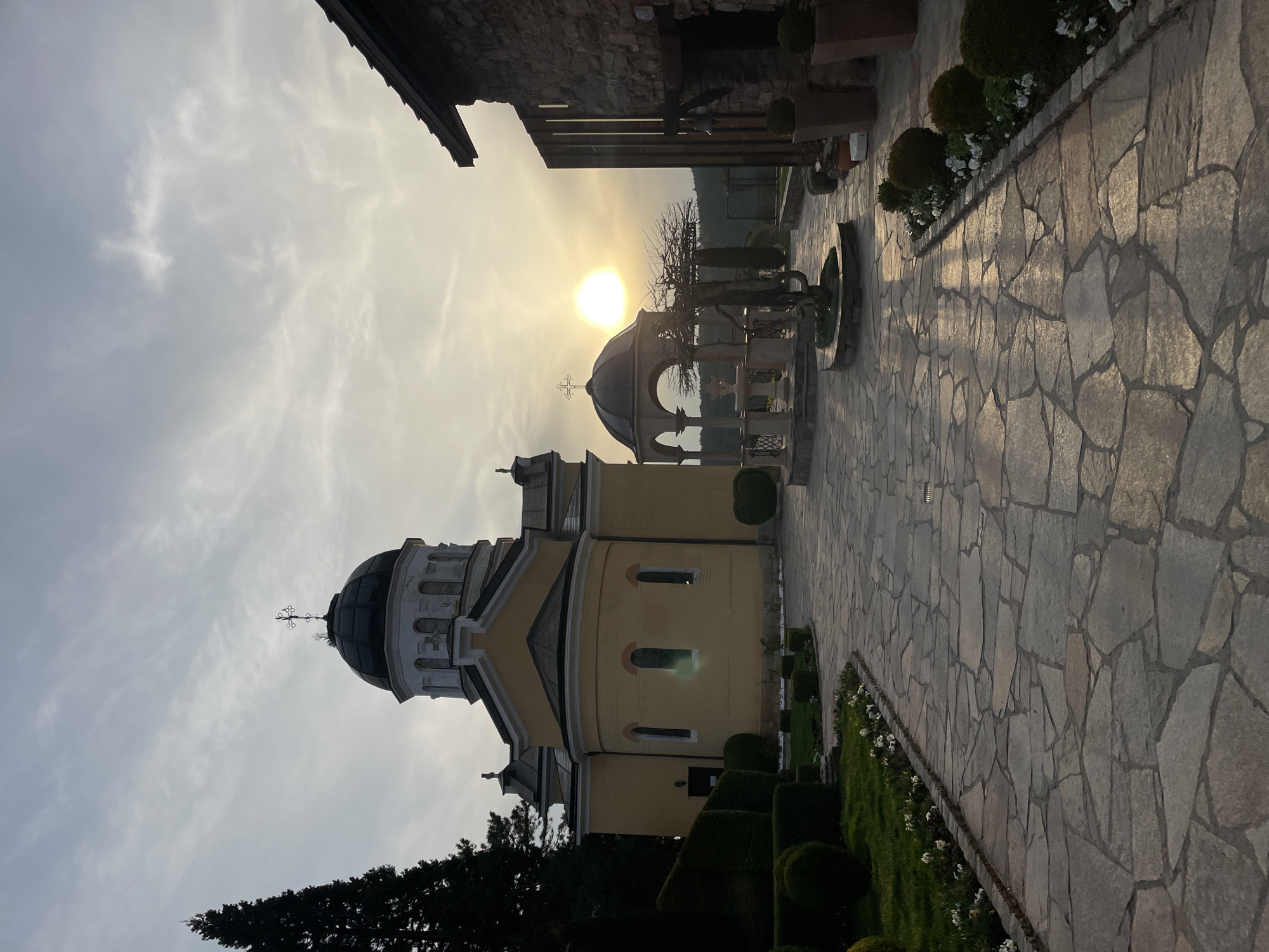 The image shows the Russian Church, also known as the Church of St. Nicholas the Miracle-Maker, located in Sofia, Bulgaria. The church has a yellow facade with white trim and a black dome topped with a cross. The sun is setting behind the church, creating a silhouette effect. There is also a stone fountain in the foreground, with a statue of Jesus on the cross atop it. To the right of the church is a stone walkway, edged with neatly trimmed bushes and white flowers. The sky is overcast.