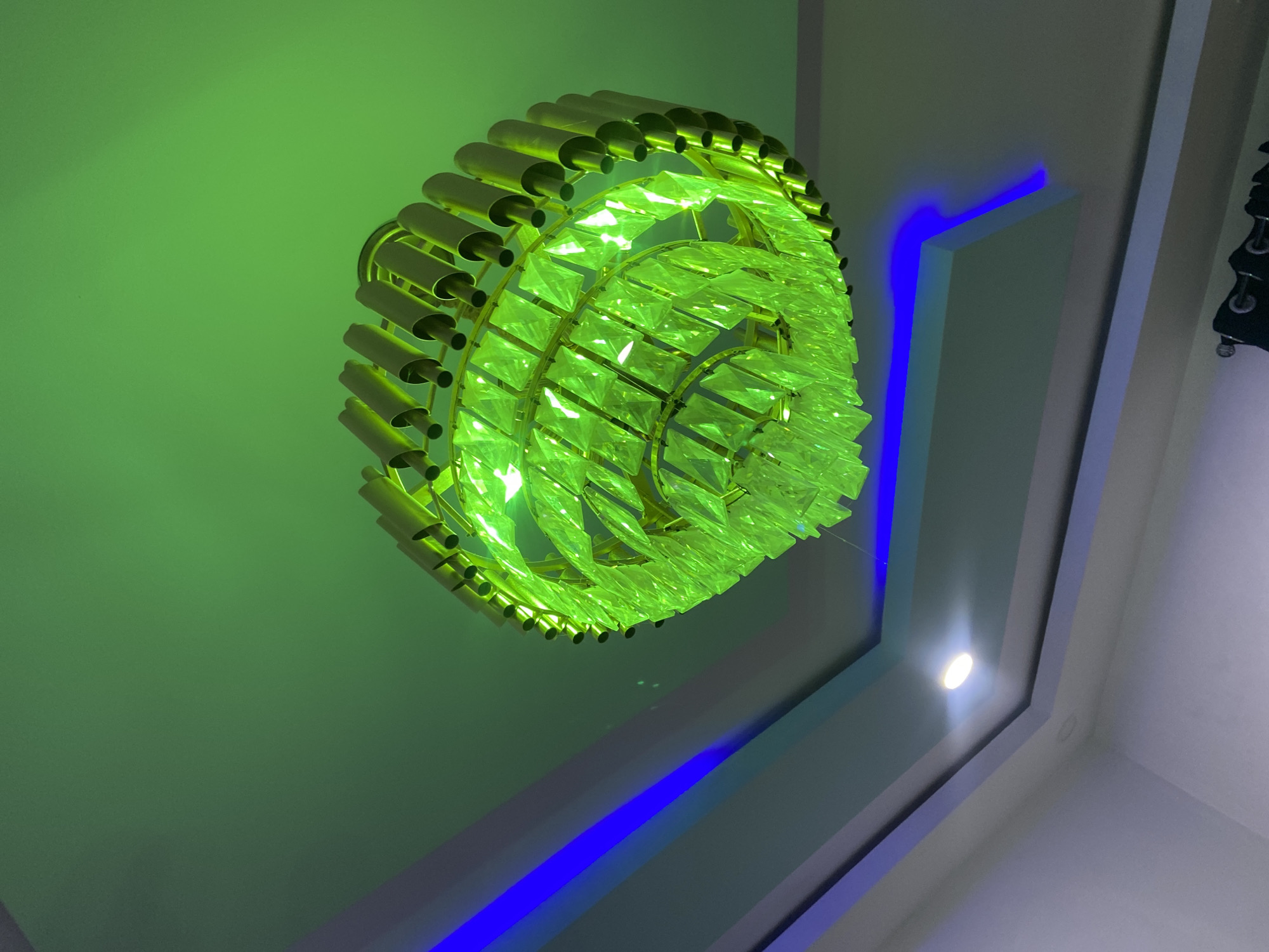 The image shows a close-up, low-angle shot of a modern chandelier with green crystal accents, suspended from a ceiling in a room. The chandelier has a metallic frame with cylindrical elements and is illuminated by green and blue LED lights embedded in the ceiling. The ceiling features a stepped design with recessed lighting. The background is a smooth, green-lit surface. The lighting creates a vibrant and contemporary ambiance.