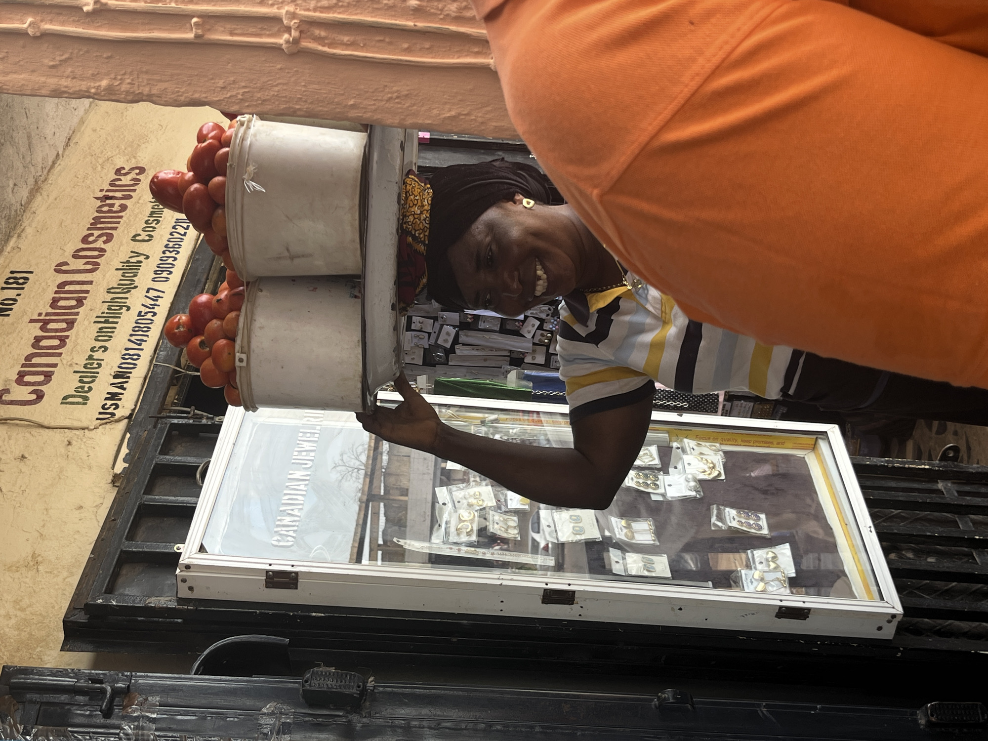 This media file captures a vibrant street scene in Jos, Nigeria, featuring a smiling woman of African descent. She is wearing a striped shirt and a headscarf, and is seen from the chest up, with her right arm extended. She is positioned in front of a display case filled with jewelry, which is labeled "CANADIAN JEWELRY". Behind her, two white buckets are filled with fresh red tomatoes. To her left, a sign reads "Canadian Cosmetics Dealers on High Quality Cosme USMAN08141805447 0909360220". The overall setting suggests an outdoor market or shop, likely during daylight hours given the illumination. The woman's joyful expression and the abundance of fresh produce and goods indicate a bustling commercial environment.