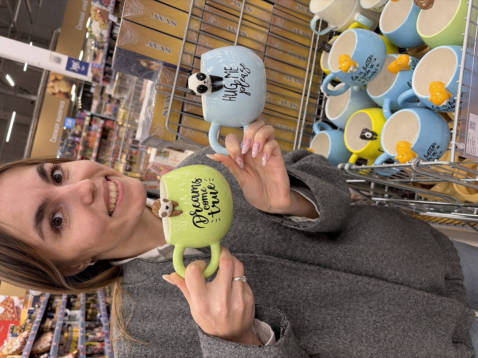 A woman holds two mugs in a supermarket aisle. The mugs are in front of a shelf stocked with similar mugs. The woman is smiling at the camera, and she is wearing a gray sweater.
The green mug she holds in her right hand has the phrase "Dreams come true" written on it. A sloth figurine is attached to the mug. The blue mug has the phrase "HUG ME please" written on it. A panda figurine is attached to that mug.
The shelf behind the woman is stocked with mugs that have various animal figurines attached to them, including dogs, sloths, and pandas. The mugs are different colors, including blue, yellow, and green. The sign on the shelf reads “Кружка HOMECLUB,” (Mug HOMECLUB) “Funny animals. ”
The shelves in the supermarket aisle are stocked with other products, including boxes of pastries. The sign on a nearby shelf says “Свежая выпечка” (Fresh baking).
The scene appears to be taken in the supermarket in the city of Izhevsk, Russia.