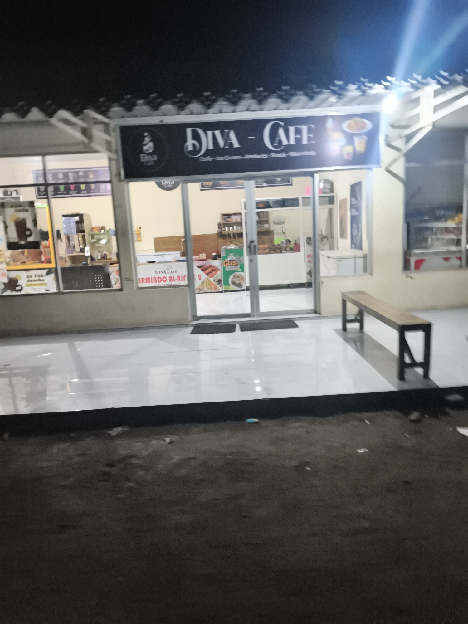 The image shows the exterior of a cafe named "Diva Cafe" at night. The cafe has a large sign with the name and pictures of food and drinks. The entrance is a glass door with reflections of the interior. Inside, shelves with products and some tables are visible. A wooden bench is placed on the tiled sidewalk in front of the cafe. The overall environment appears to be a street in Ketawang, Indonesia, with a somewhat unlit and dark setting typical of a nighttime street scene.