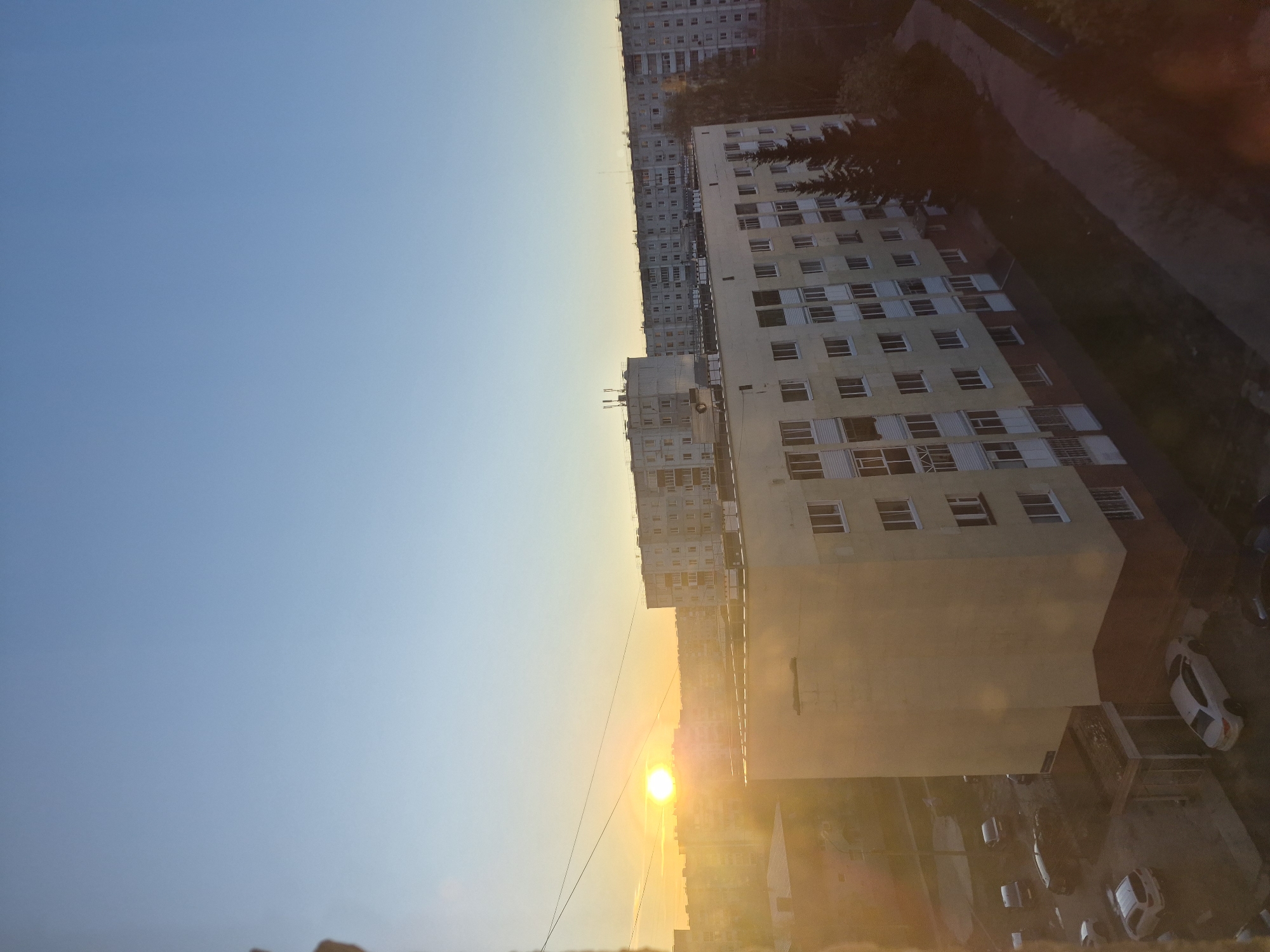 A street scene in Tobolsk, Russia, features a sunrise partially obscured by buildings. The sky transitions from a clear blue at the top to a yellow-orange hue near the horizon, where the sun is visible. Several apartment buildings are in the frame, with the closest one a light yellow color and multiple windows. Below, parked cars are visible on the street.
