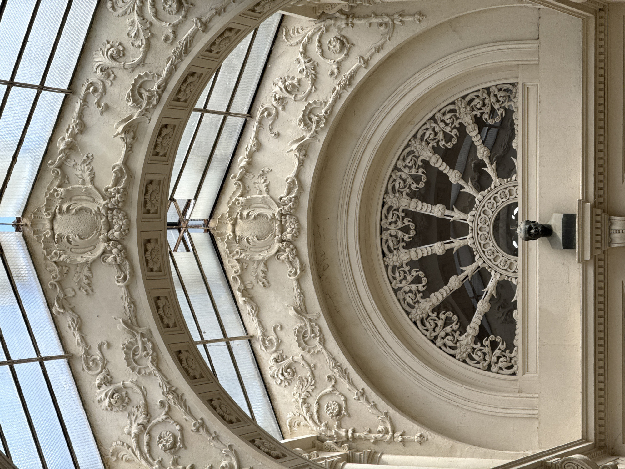 The image shows an ornate architectural detail with a semi-circular window, featuring intricate plasterwork. Above the window, decorative plaster mouldings include scrolling acanthus leaves, floral motifs, and cartouches. The window itself has a radial pattern of ornamental metalwork. A bust of a man is visible below the window. The structure appears to be an interior atrium or covered walkway, with a glass roof and metal framing visible at the top. The building is located in Nantes, France, and is part of the Passage Pommeraye.