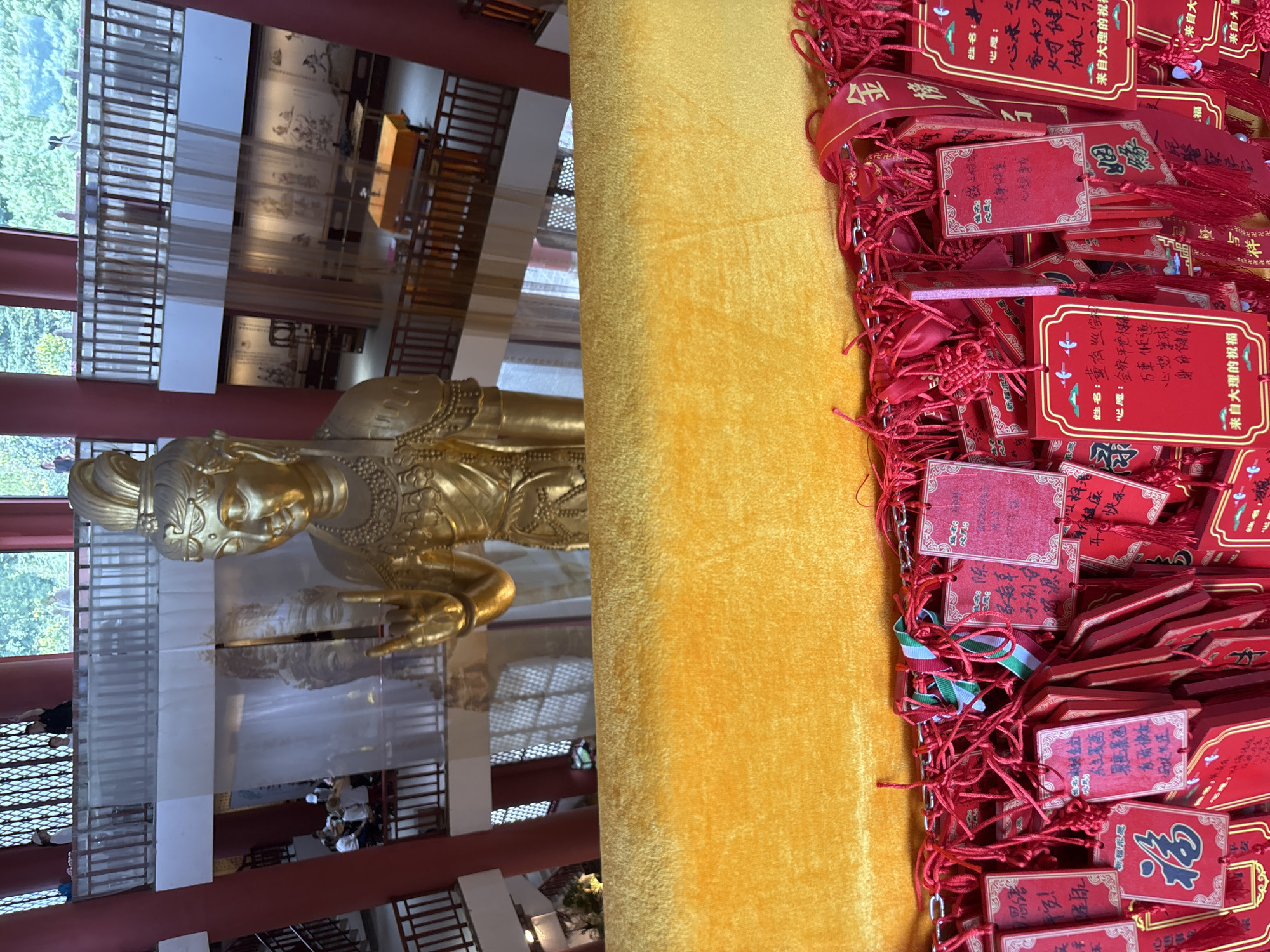 The image depicts the interior of a multi-level building, likely a temple or a tourist attraction, in Dali City, China. In the foreground, a large, golden statue of a deity, possibly Guanyin, is partially visible from the chest up. The deity is adorned with intricate jewelry and is making a hand gesture.
Dominating the lower part of the image is a vibrant yellow, velvety fabric draped over a railing. Hanging from a metal chain above this fabric are numerous small, red tags. These tags, adorned with Chinese characters and tassels, appear to be prayer requests or good luck charms. Some of these tags have red ribbons attached, with some ribbons featuring gold Chinese characters.
The background reveals a series of balconies and walkways with wooden railings. Several individuals are visible on these upper levels, observing the scene below or interacting. One person on an upper balcony is pointing upwards. The architecture suggests a traditional Chinese style. Further in the background, a large, sheer white curtain is visible, behind which another, more faded statue or artwork can be discerned. More traditional paintings or murals are displayed on the walls of the lower levels, and some orange-covered tables are present. The overall environment is well-lit, with natural light filtering in from above and possibly windows.