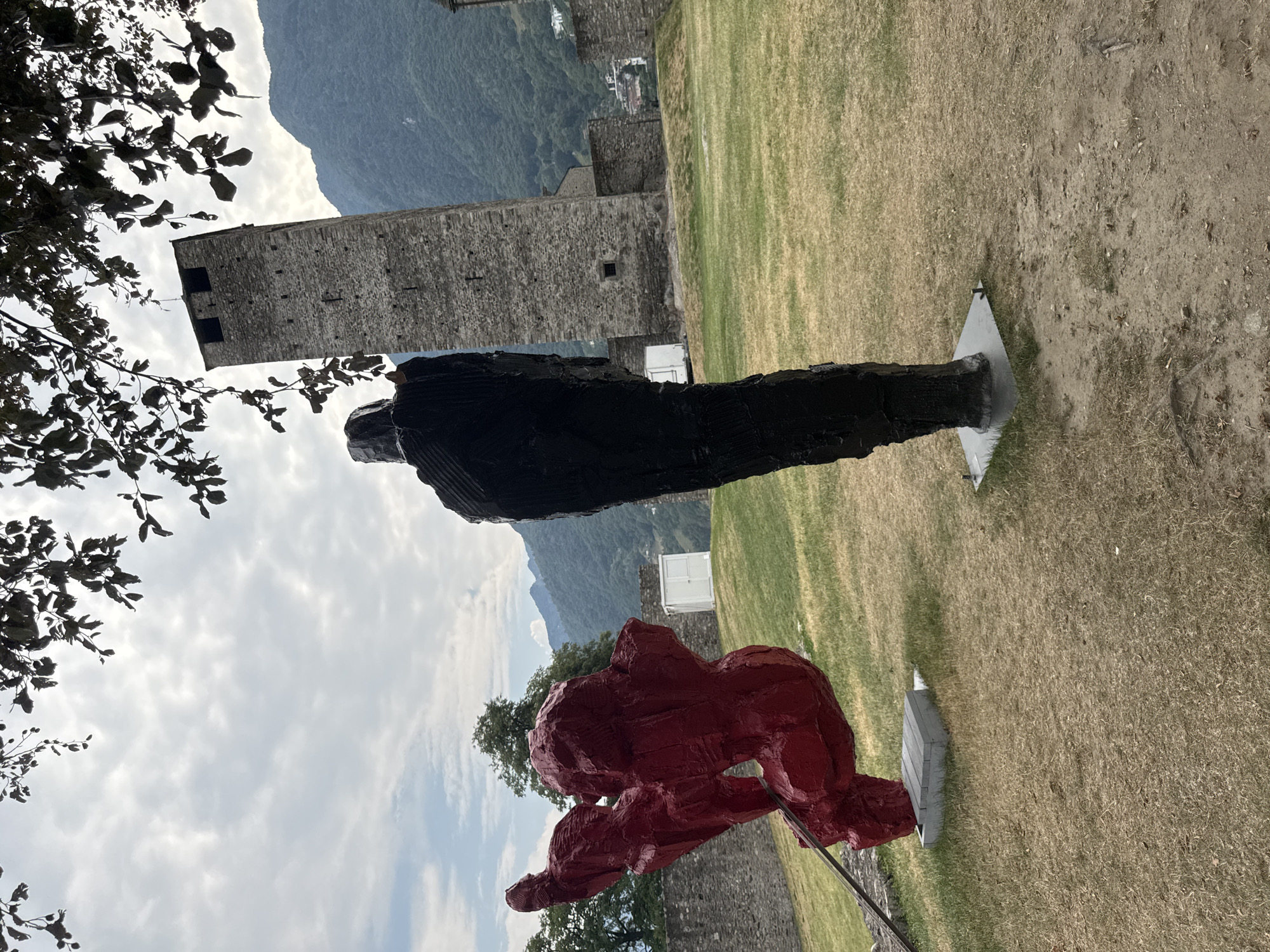 The image depicts a scene at Castel Grande in Bellinzona, Switzerland. In the foreground, two modern sculptures are visible on a grassy lawn. The sculpture on the left is abstract and bright red, with a dynamic, upward-reaching form. The sculpture on the right is a tall, dark, textured figure, standing upright. Behind the sculptures, a section of ancient stone wall, part of the Castel Grande complex, runs horizontally across the middle ground. Further back, a prominent stone tower, characteristic of Castel Grande, rises vertically, its weathered stone facade contrasting with the cloudy sky. The background features rolling green mountains and a small town nestled in the valley. The overall atmosphere is one of historical architecture blended with contemporary art in an outdoor setting.