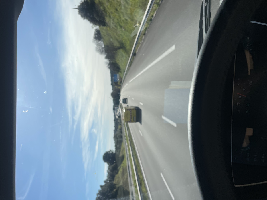 The image displays a daytime scene from inside a vehicle traveling on a multi-lane highway in Tavel, France. The top portion of a dark steering wheel and a dashboard speedometer, showing a speed of approximately 82 units (likely km/h), are present at the bottom of the frame. The asphalt road features white dashed lane markings and extends into the distance. A bright yellow utility van is positioned in the middle lane, moving away from the camera, with a dark passenger vehicle visible further ahead. Guardrails line the right side of the highway, beyond which are vegetated slopes covered with green grass and various trees and shrubs. The sky is clear blue with scattered thin white clouds. A blue road sign is partially visible on the distant right side of the highway.