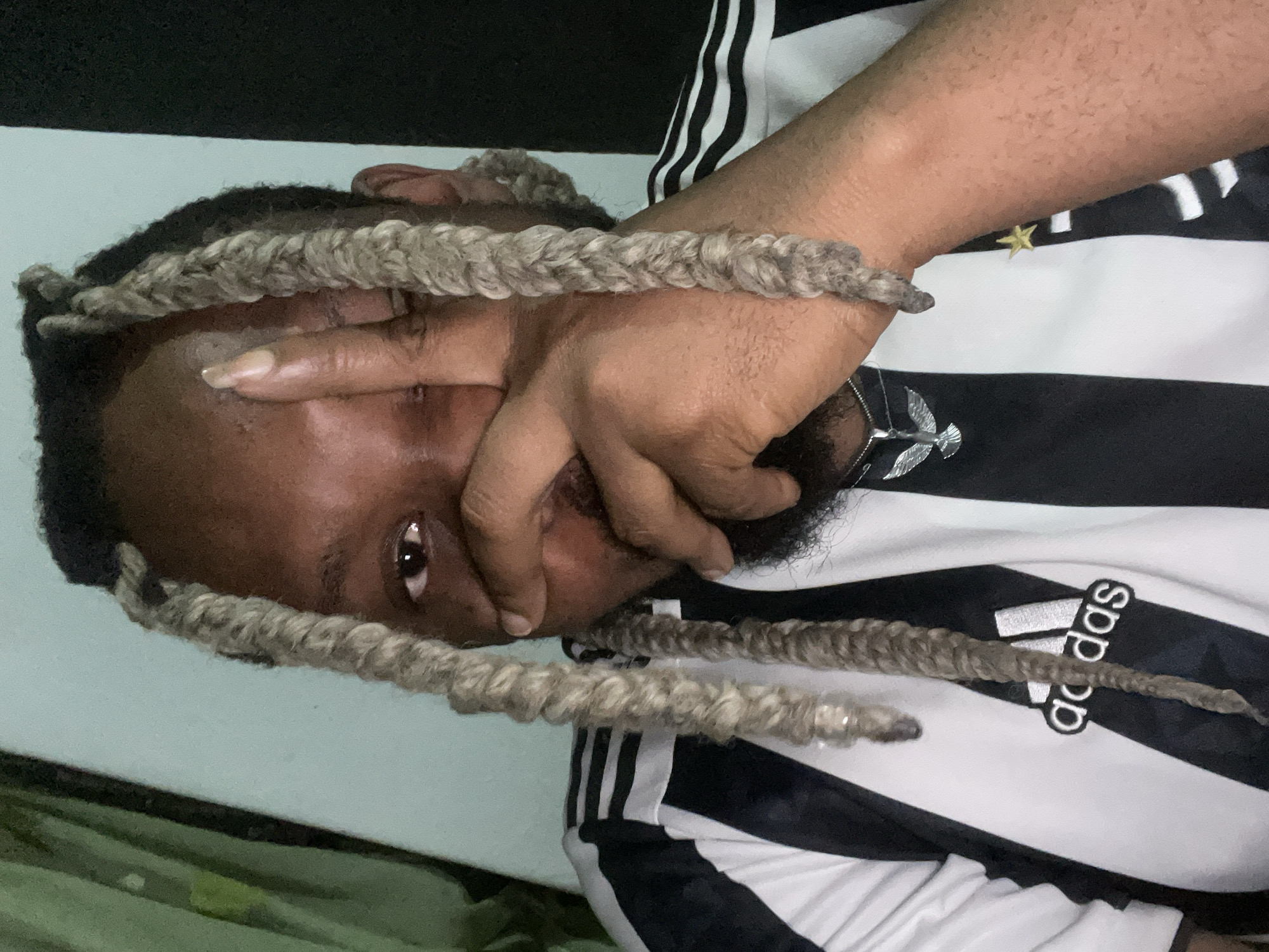 The image shows a close-up of a Black person wearing a black and white Adidas jersey. Their face is partially obscured by their right hand, which is held up in front of their eye. The person has long, light-colored dreadlocks. A silver necklace with a bird pendant is visible on their chest. The background is a light blue wall. The image does not contain any identifiable landmarks or contextual information that would allow for the determination of its location within the city of Jos, Nigeria.