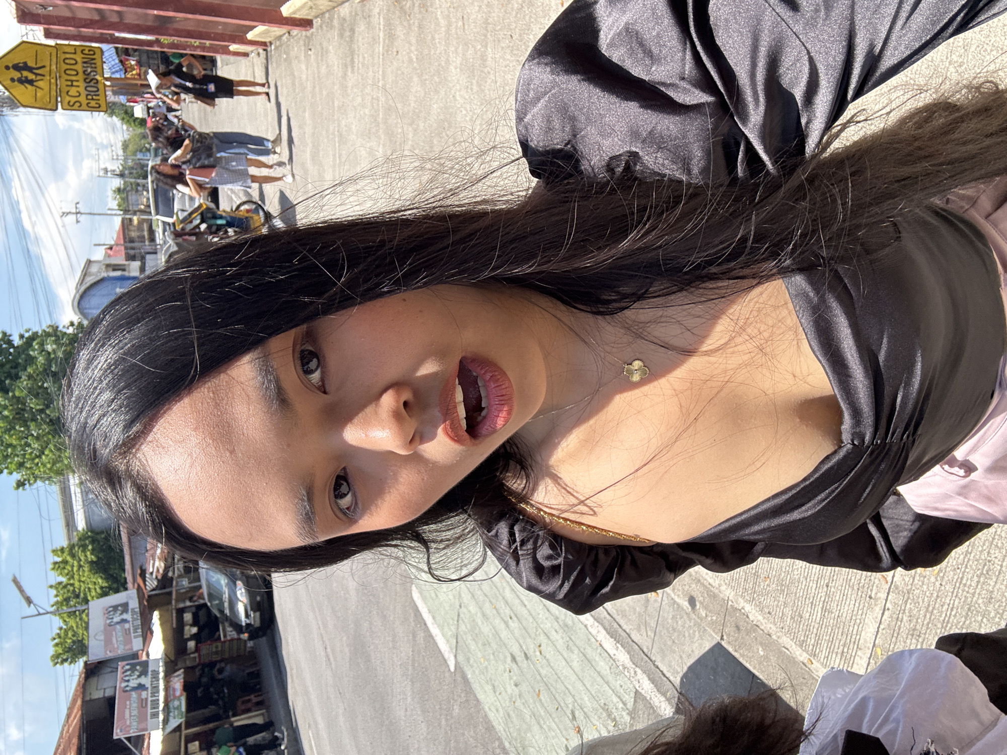 The image captures a selfie taken by a young woman against the backdrop of a bustling street scene in Davao City, Philippines. The main subject, with long, straight dark hair, is positioned prominently in the foreground, looking directly at the camera. She wears a black, satiny top with puffy sleeves and has a slightly open-mouthed expression with her lips tinted. A delicate gold necklace with a four-leaf clover pendant adorns her neck.
The background reveals a sunny outdoor setting with strong shadows, indicating bright daylight. A concrete sidewalk lines the left side of the frame, alongside an asphalt road. Several other people are visible in the mid-ground, walking along the street.
Notable objects include a yellow, diamond-shaped "SCHOOL CROSSING" sign with black lettering and pedestrian symbols. Further down the street, various small establishments or vendor stalls with corrugated iron roofs and hanging banners are present. While most text on these banners is illegible, words like "INUMAN" and "PARES MAMI" can be faintly discerned on one, suggesting a food or drink business. A dark-colored vehicle and parts of a bicycle or motorbike are also visible on the street. The sky above is clear blue with some scattered white clouds, and green tree foliage adds to the outdoor ambiance. The scene depicts a lively, everyday urban environment.