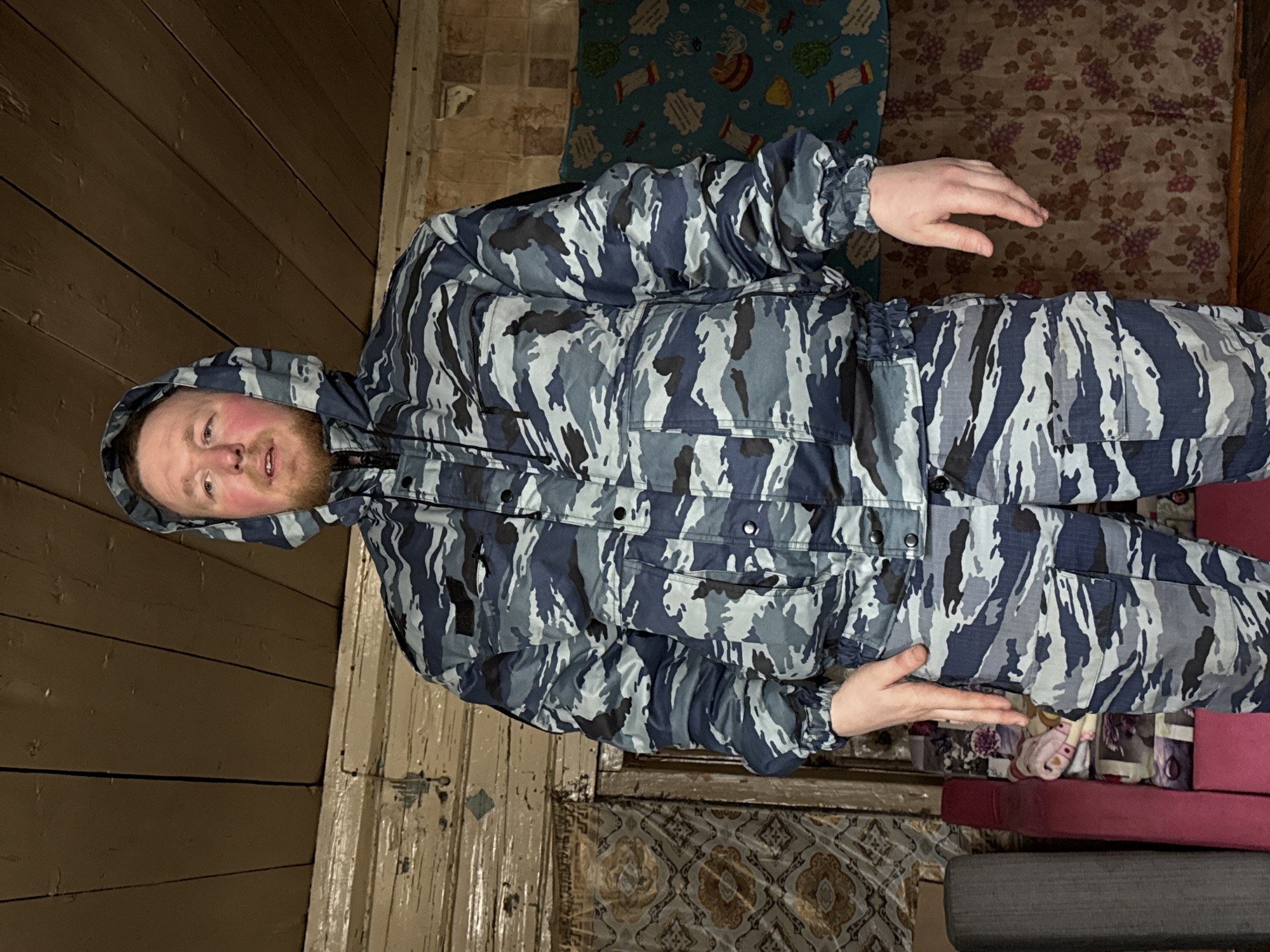 A man is standing indoors, wearing a blue and gray camouflage jacket with a hood up. The background includes wooden walls and various patterned carpets or blankets. A portion of stacked cushions is visible near the bottom right. The man's location is Nizhnyaya Chermoda, Russia.