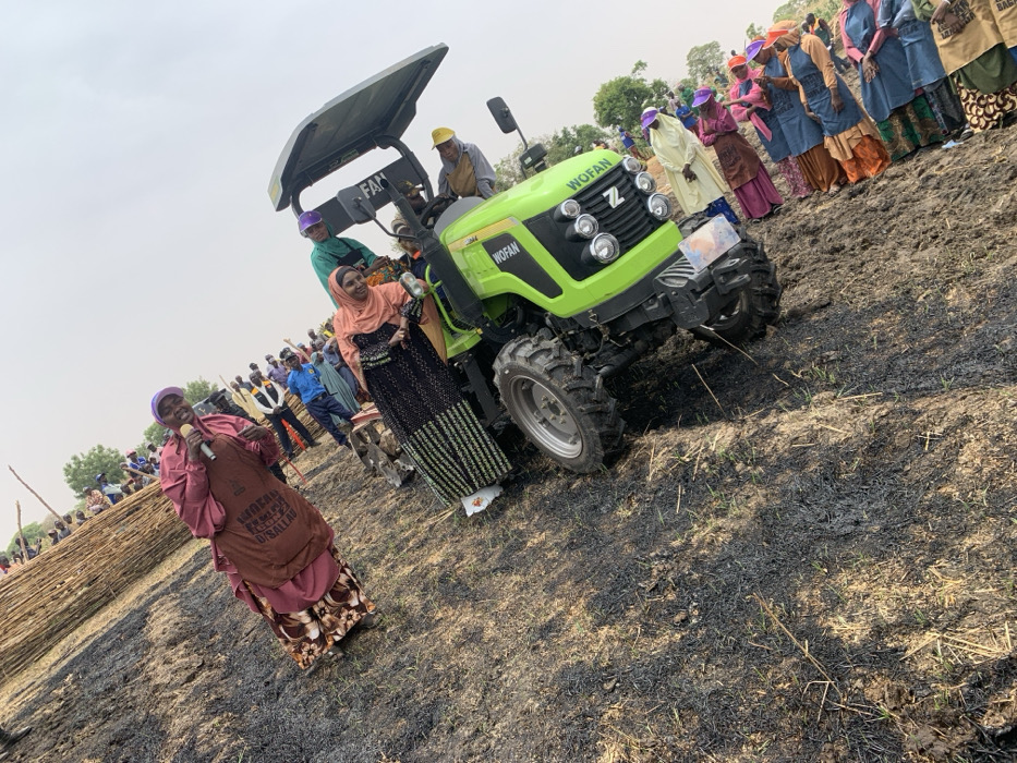 The scene is a gathering of people in Ringimawa, Nigeria, located at the coordinates 11.666 latitude, 8.414 longitude. There are several people present, both men and women. The people are standing in front of a lime green tractor, while others are seated on the tractor. The tractor is brand new. The people are dressed in various colorful clothing. The ground is covered in black burnt grass, possibly the aftermath of a recent fire. One person is holding a microphone and addressing the group. The overall atmosphere is one of celebration and excitement.