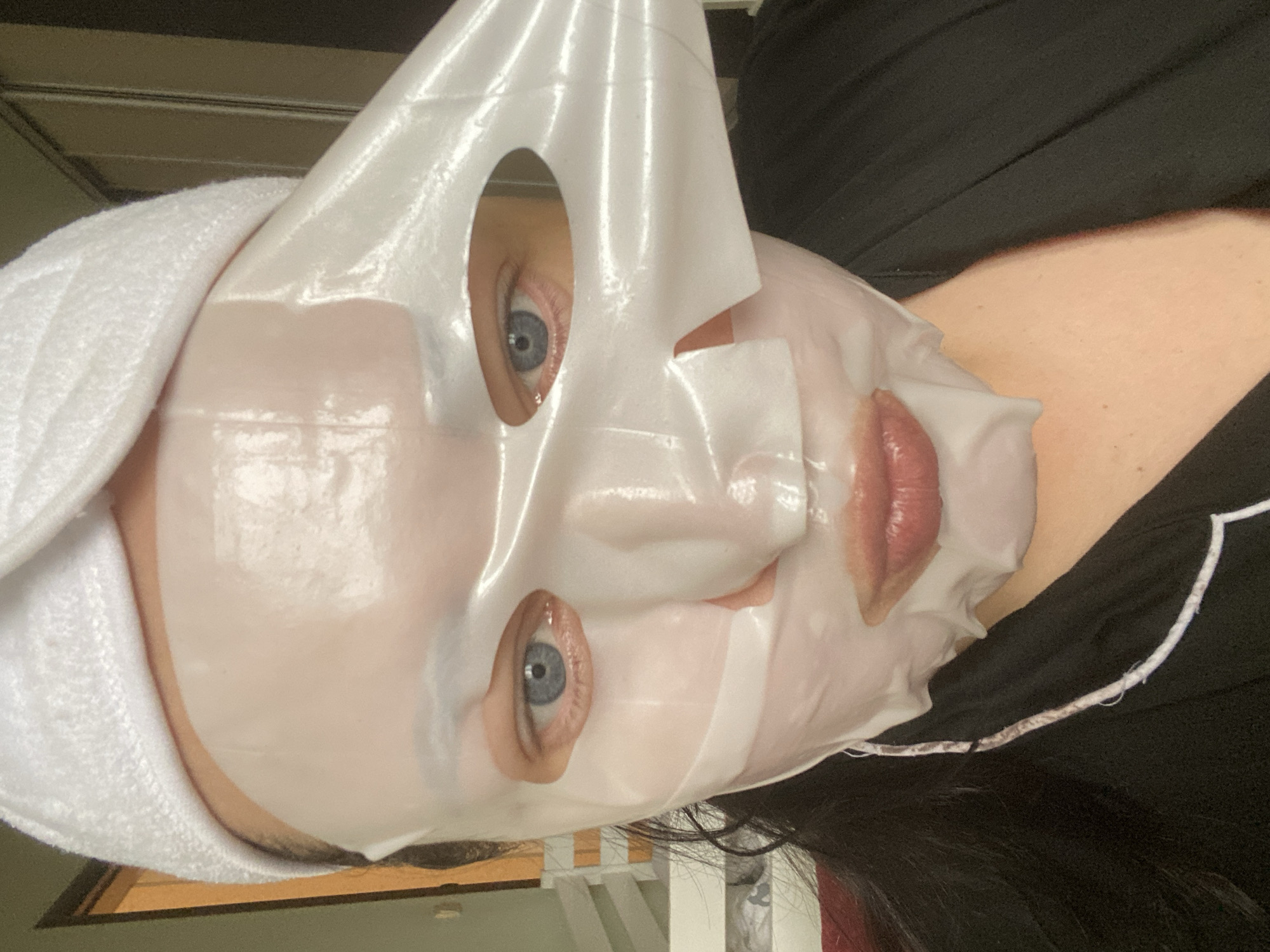 A person is wearing a sheet mask and a white towel wrapped around their head. The mask has openings for the eyes and mouth. The person has light skin and blue eyes. They are wearing a black top with white piping. The background is blurry and indistinct, but appears to be an indoor setting with a muted color palette. There is no evidence in the image to suggest that it was taken in Sochi, Russia.