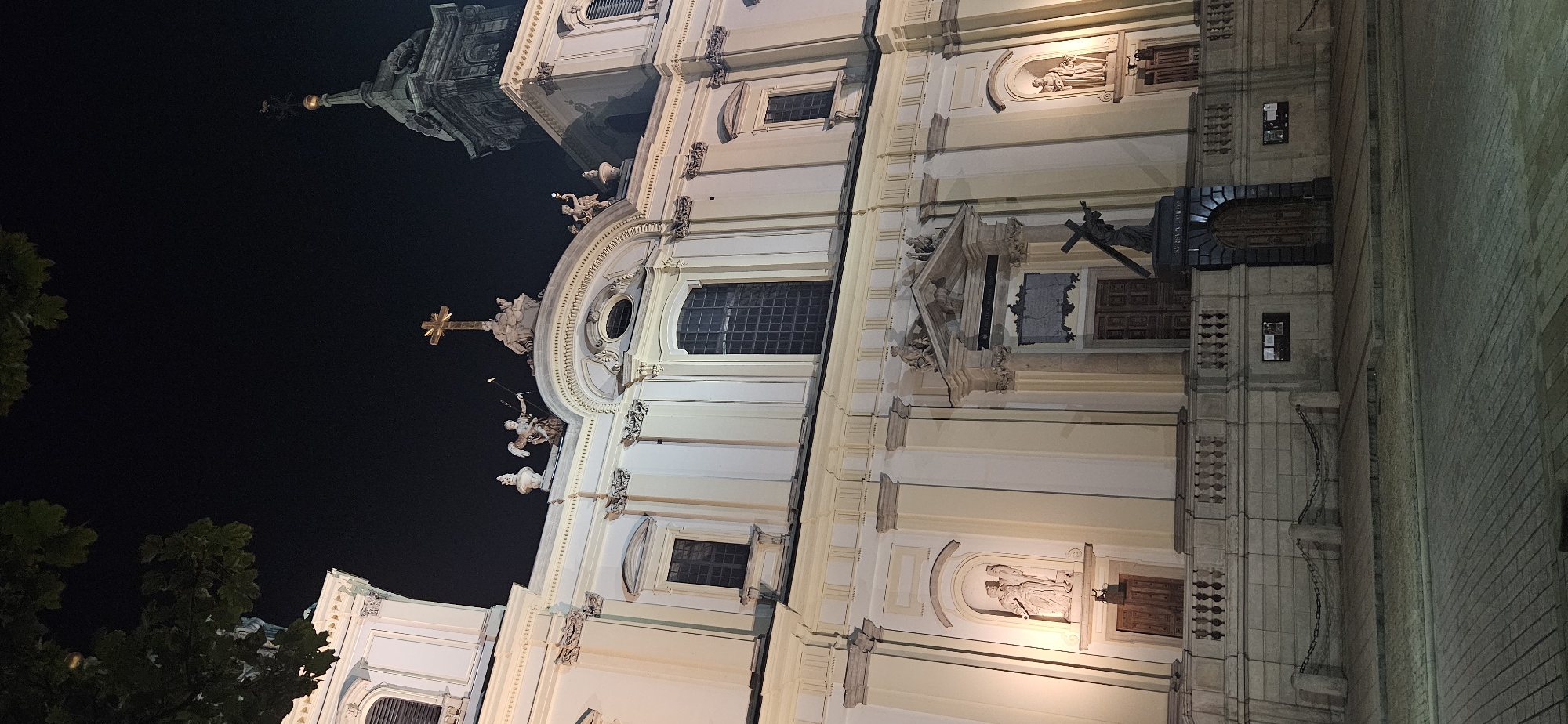 The image shows the facade of a large, ornate church at night in Warsaw, Poland. The building is illuminated by warm lights, highlighting its classical architecture with multiple columns, arched windows, and decorative statues. A prominent golden cross is visible at the top of the central pediment, and a statue of a figure carrying a cross stands in front of the main entrance. The foreground shows a cobblestone street and a stone balustrade, with chains marking off a pathway. Trees partially obscure the top left corner of the image.