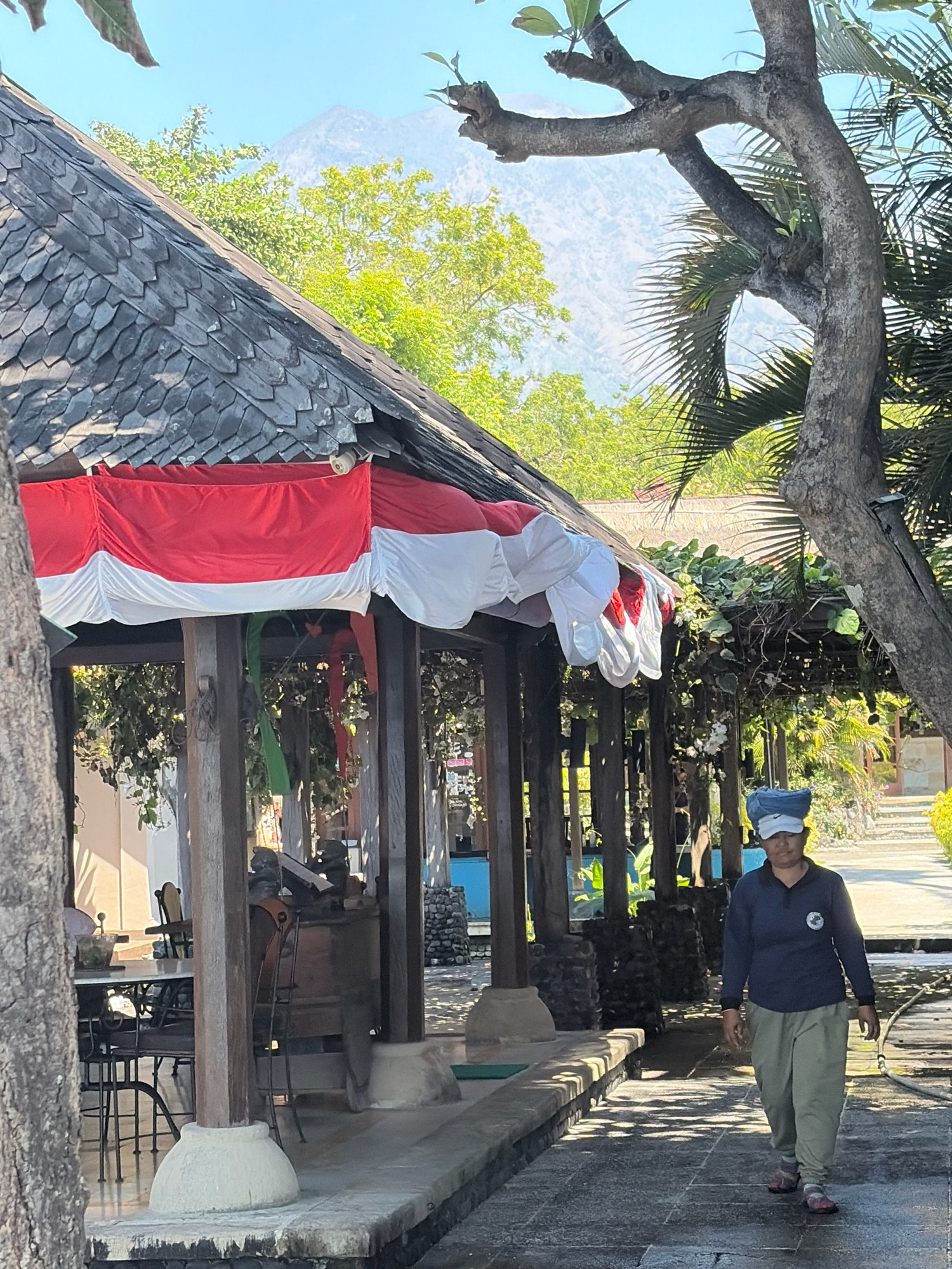A medium-shot image captures an outdoor scene in Kubu, Indonesia, with a focus on a woman walking on a paved pathway. The woman, wearing a blue long-sleeved shirt with a circular emblem on the left chest, light olive-green pants, a blue headscarf, and a white baseball cap, is walking from right to left towards the foreground. Her face is partially obscured by her headwear.
Behind her, a traditional Balinese structure with a tiled, multi-layered roof is visible. Red and white fabric, resembling a flag or banner, is draped along the front edge of the structure. The structure is supported by wooden pillars resting on stone bases. In the background, lush green trees and vegetation are present, with a hazy mountain peak visible in the distance under a clear blue sky. Sunlight casts shadows on the paved pathway, indicating a bright day. The environment appears to be a resort or a public area with a pathway and seating areas partially visible to the left.