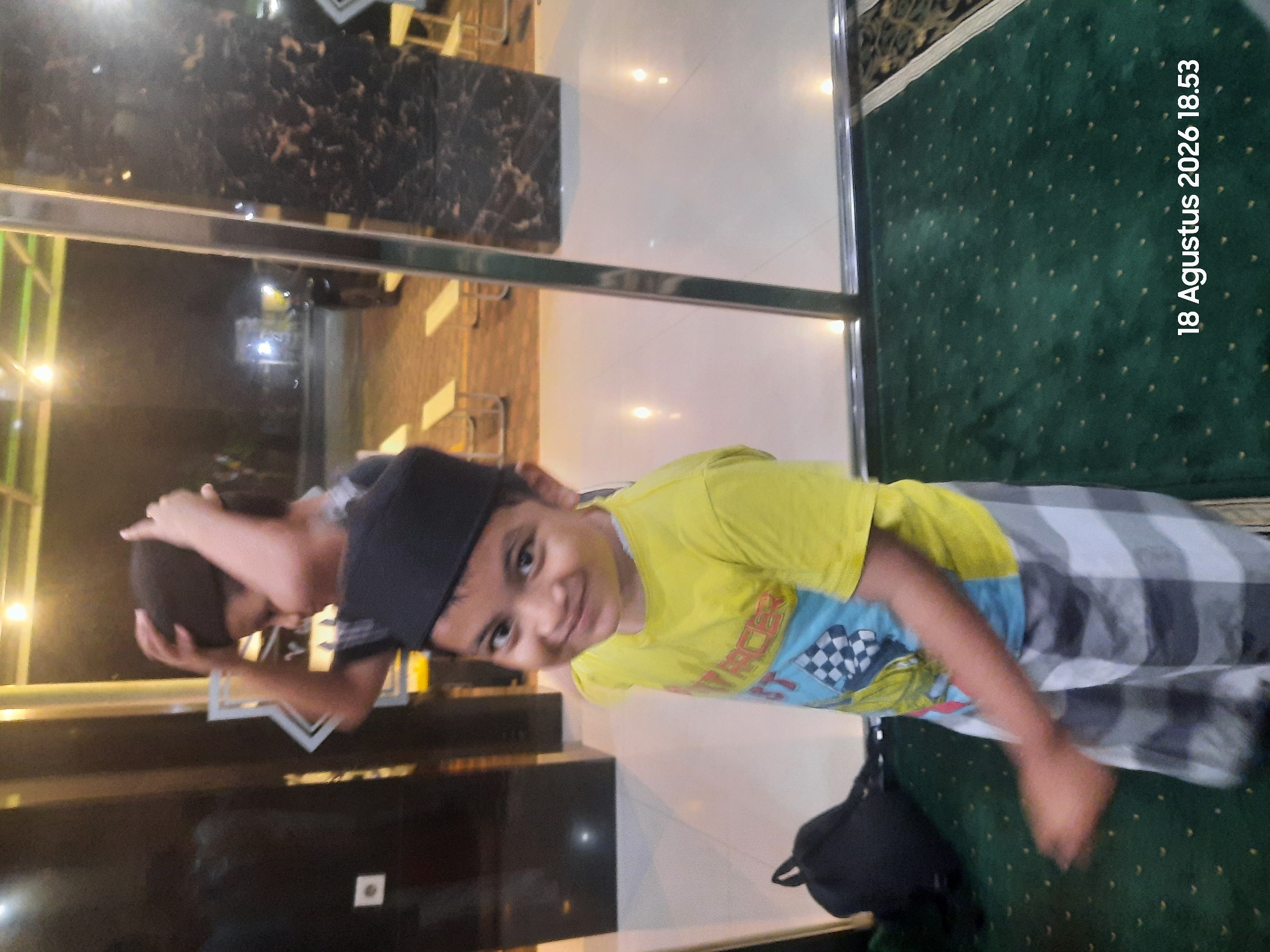 In the city of Sukokerto, Indonesia, a young boy, estimated to be between five and seven years old, is the primary subject of the image. He is wearing a yellow t-shirt with a checkered flag graphic and a black kufi, and plaid shorts. He is smiling and looking at the camera. Behind him, another child is visible, with their hands on their head. The scene appears to be indoors, possibly a community center or a public gathering space, with a large dark marble pillar and glass doors. The floor is tiled and a green carpet with a pattern of small dots is present in the foreground. A black backpack is on the floor to the left. The lighting suggests it is evening. The timestamp "18 Agustus 2026 18.53" is superimposed on the image.