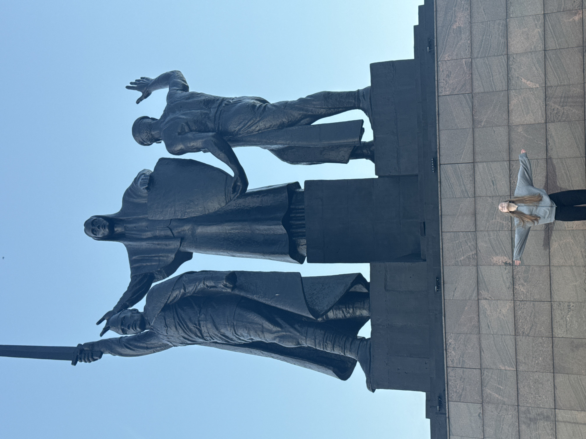 The photograph captures a low-angle view of a monumental bronze sculpture depicting three figures against a clear blue sky. The sculpture is situated in the city of Perm, Russia, specifically at the Perm-1 railway station.
In the center of the sculpture stands a female figure with outstretched arms, holding a shield. To her left, a male figure in military attire raises a sword. To her right, another male figure, likely a worker, has his right arm raised in a gesture of greeting or affirmation. The figures are positioned on a multi-tiered dark pedestal, which is itself mounted on a large, granite-clad plinth.
In the foreground, a young woman with long, light-brown hair is standing with her arms extended to her sides, mirroring the gesture of the central female figure in the sculpture. She is wearing a gray hooded sweatshirt and dark pants. The woman is positioned at the base of the granite plinth, emphasizing the scale of the monument. The overall lighting suggests it is daytime, likely morning or late afternoon, with soft shadows. The environment appears to be an urban outdoor setting, indicated by the man-made structures of the sculpture and plinth.