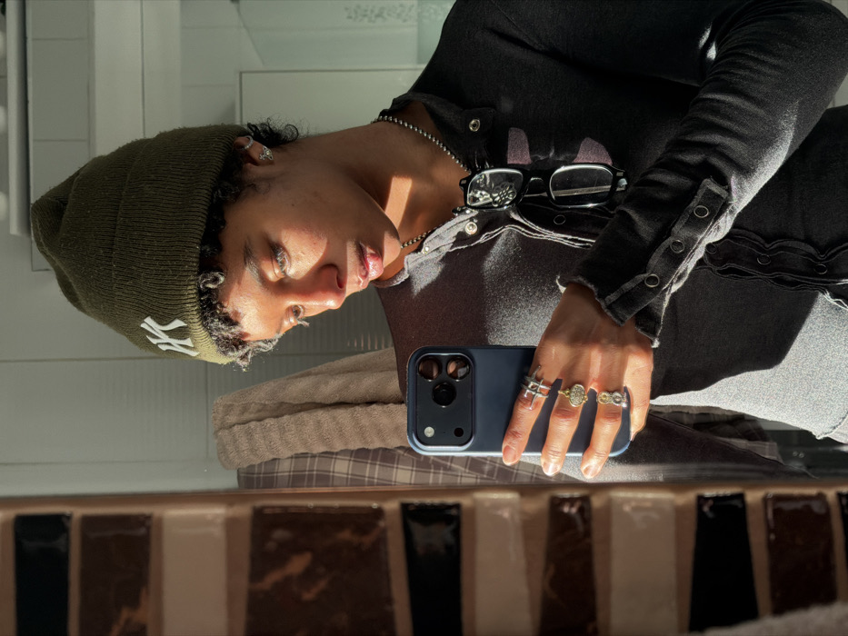 An individual, appearing female, is depicted taking a mirror selfie indoors in Champs-sur-Marne, France. The subject wears an olive green beanie with a white "NY" logo and a dark gray long-sleeved top with snap buttons and distressed edges. A silver-colored beaded necklace holds a pair of black-framed eyeglasses. A silver-colored stud earring is visible in the right ear. The subject's left hand holds a dark blue smartphone, angled for the selfie, and wears multiple rings: a silver-colored interwoven ring on the index finger, a gold-colored ornate ring on the middle finger, and two rings (one gold, one silver) on the ring finger. Strong natural sunlight illuminates the right side of the subject's face, highlighting light-colored eyes. The background, reflected in the mirror, shows a section of a tiled wall with brown, black, and light-colored rectangular tiles in a staggered pattern, and a light-colored, textured towel hanging above the tiles.