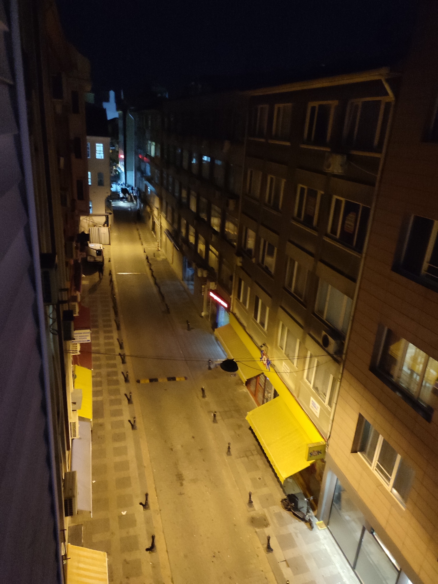 This is a nighttime, aerial shot of a narrow street in Kadıköy, Turkey, likely a commercial area with apartment buildings lining both sides. The street is paved with cobblestones and illuminated by streetlights, casting long shadows. Yellow awnings extend from the ground-floor businesses on the right side of the street, one of which has a "SOK" logo visible. A parked scooter is situated beneath the awnings. On the left side of the street, the buildings are taller and have visible windows, some of which are lit. A white truck is parked further down the street, partially obscured by shadows. The sky is dark, indicating it is nighttime.