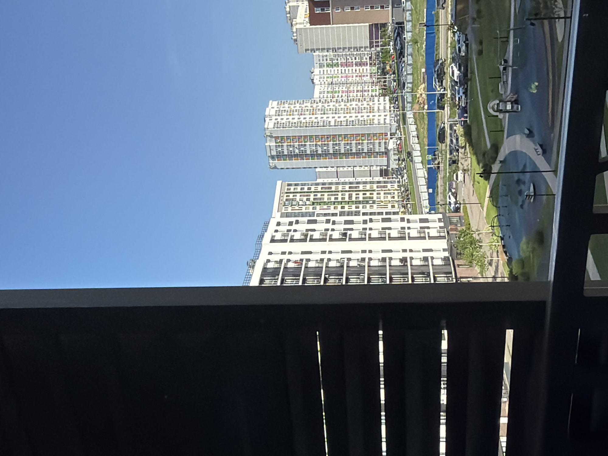 The image depicts a view from a high vantage point, likely a balcony, overlooking a cityscape in Pervomaysky, Russia. A dark, slatted structure, part of the balcony itself, dominates the foreground. Beyond this structure are multiple high-rise apartment buildings, arranged linearly in the middle ground. These buildings are predominantly white with repeating window patterns. In between the buildings, there's a strip of green space containing a playground. A parking lot, filled with cars, runs along one side of the playground. The sky above is clear and blue.
